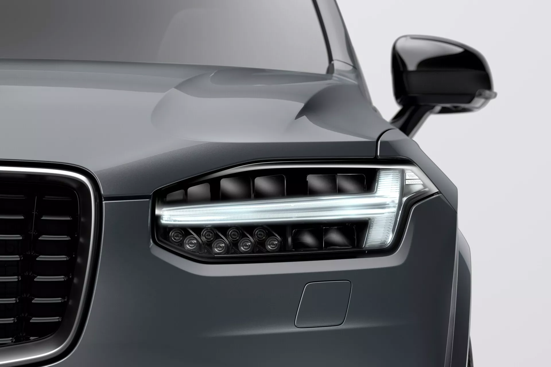 Zbliżenie przedniego lewego rogu Volvo XC90 z 2015 roku (2. generacja) pokazujące nowoczesny design reflektorów LED z kątowymi światłami do jazdy dziennej, częściowy widok dużej kratki z pionowym wzorem, błyszczący ciemnoszary metalik i czujnik parkowania na zderzaku, wzięty z niskiego kąta od prawej do lewej.