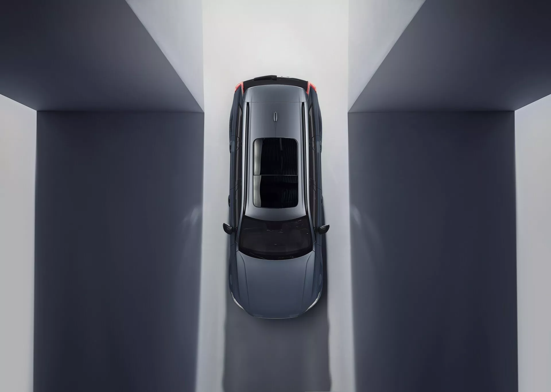 Widok z lotu ptaka na Volvo XC90 2015 (2. generacja) ukazujący symetryczną kompozycję i odbicia środowiskowe. Centralne położenie w środowisku wewnętrznym o neutralnych tonach podkreśla dach samochodu z szyberdachem lub panoramicznym szklanym dachem. Widoczne części obejmują maskę, dach, bagażnik i górną połowę drzwi, podkreślając eleganckie linie i krzywizny sugerujące luksusowy design skoncentrowany na aerodynamice i przestronnym oświetleniu wnętrza.