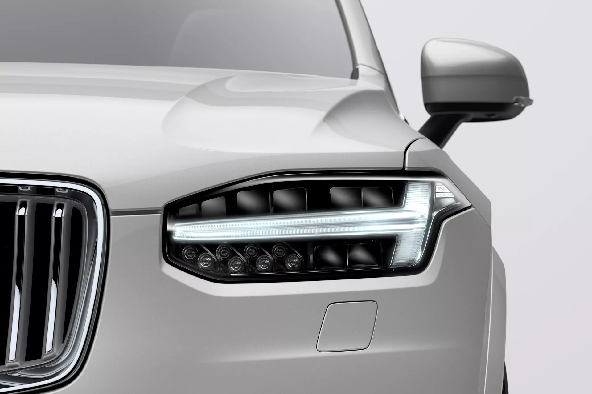 Zbliżenie na przedni lewy reflektor i część atrapy chłodnicy Volvo XC90 z 2015 roku (druga generacja). Elegancki reflektor integruje wydłużone, zaawansowane elementy LED/HID z przezroczystą soczewką, prezentując kanciasty, aerodynamiczny design. Przylegająca chromowana kratka z pionowymi żaluzjami podkreśla nowoczesny, luksusowy wygląd pojazdu. Panel nadwozia, prawdopodobnie w neutralnym kolorze, takim jak srebrny, pokazuje płynne przejście od reflektora. Częściowo widoczne jest również lusterko boczne. Zdjęcie podkreśla współczesny styl samochodu i zaawansowaną technologię oświetlenia.
