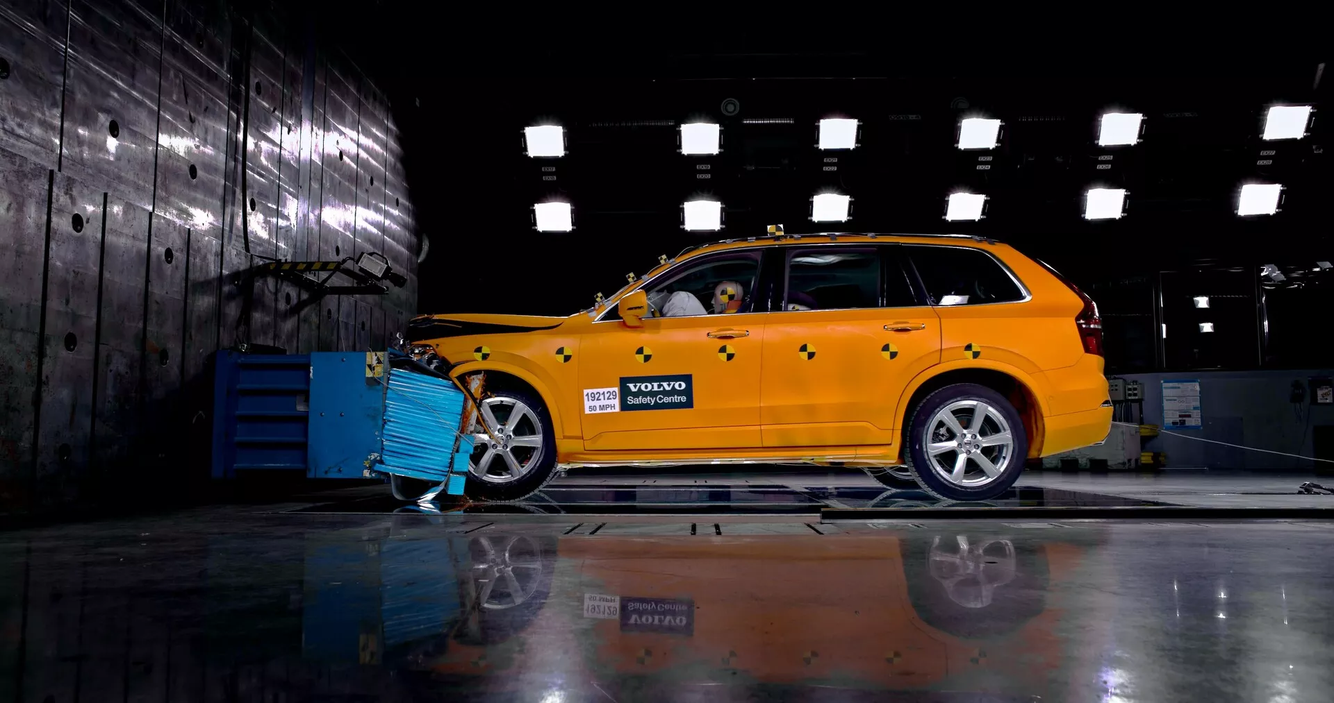 Widok boczny jasnożółtego Volvo XC90 z 2015 roku (2. generacja) w przemysłowym otoczeniu, prezentujący jego design kombi z widoczną przednią kratką, reflektorami i felgami ze stopów lekkich. Samochód jest podłączony do niebieskiego urządzenia diagnostycznego lub ładowarki, a manekin wewnątrz sugeruje demonstrację testu zderzeniowego lub bezpieczeństwa. Polerowana podłoga odbija podwozie i koła po przeciwnej stronie.