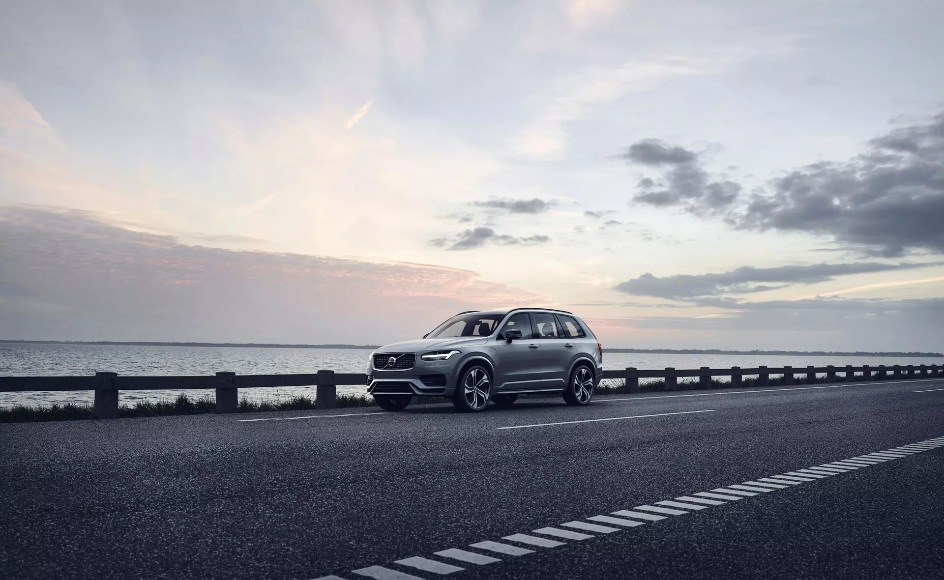 Nowoczesny SUV, konkretnie Volvo XC90 z 2015 roku (2. generacja), zaparkowany po prawej stronie dwupasmowej drogi. Pojazd uchwycony jest pod kątem trzech czwartych przodu, ukazując przedni grill, reflektory, maskę i błotnik po stronie kierowcy. Kąt ten ujawnia również część profilu samochodu, w tym koła, drzwi kierowcy i część tyłu. Koła są lekko skręcone w prawo, z potencjalnie sportowymi elementami designu, takimi jak wyraźny zderzak i większe wloty powietrza, co sugeruje dynamiczny pakiet. Zdjęcie, prawdopodobnie wykonane o zmierzchu przy miękkim, rozproszonym świetle pod lekko pochmurnym niebem, podkreśla spokojną atmosferę. Dobrze zdefiniowana karoseria z widocznymi liniami charakteru wskazuje na luksusowy model. Wnioskując z obrazu, cechy technologiczne obejmują reflektory LED, zaawansowane wzory kół, wyrafinowaną przednią kratkę z możliwymi czujnikami lub kamerami oraz aerodynamiczne akcenty. To wysokiej jakości zdjęcie może być odpowiednie do celów reklamowych, podkreślając samochód jako luksusowy wybór stylu życia.