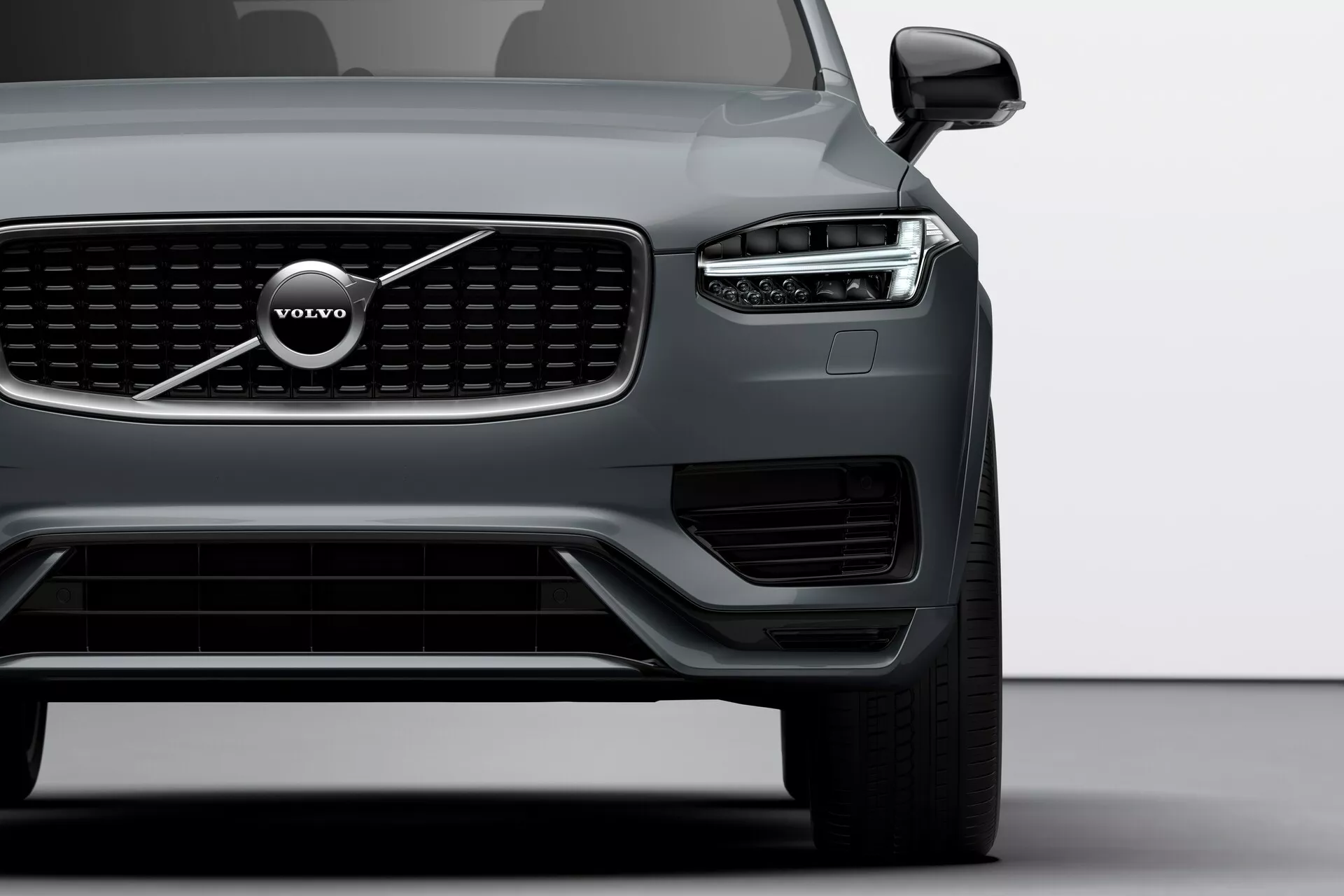 Widok ćwiartki przedniej lewej części Volvo XC90 z 2015 roku (2. generacja), uchwycony pod niskim kątem, ukazujący ostre, kątowe reflektory z charakterystycznymi diodami LED z dziennymi światłami w kształcie młota Thora, wyróżniającą się kratkę przednią pionowymi listwami i emblematem Volvo, szerokim dolnym wlotem powietrza z dodatkowymi światłami/sensorami, aerodynamiczne kontury maski, eleganckie lusterko zewnętrzne z zintegrowanym kierunkowskazem, częściowo widoczne duże wieloramienne aluminiowe koło i matowe wykończenie nadwozia, podkreślające nowoczesność design i zaawansowaną technologię pojazdu.