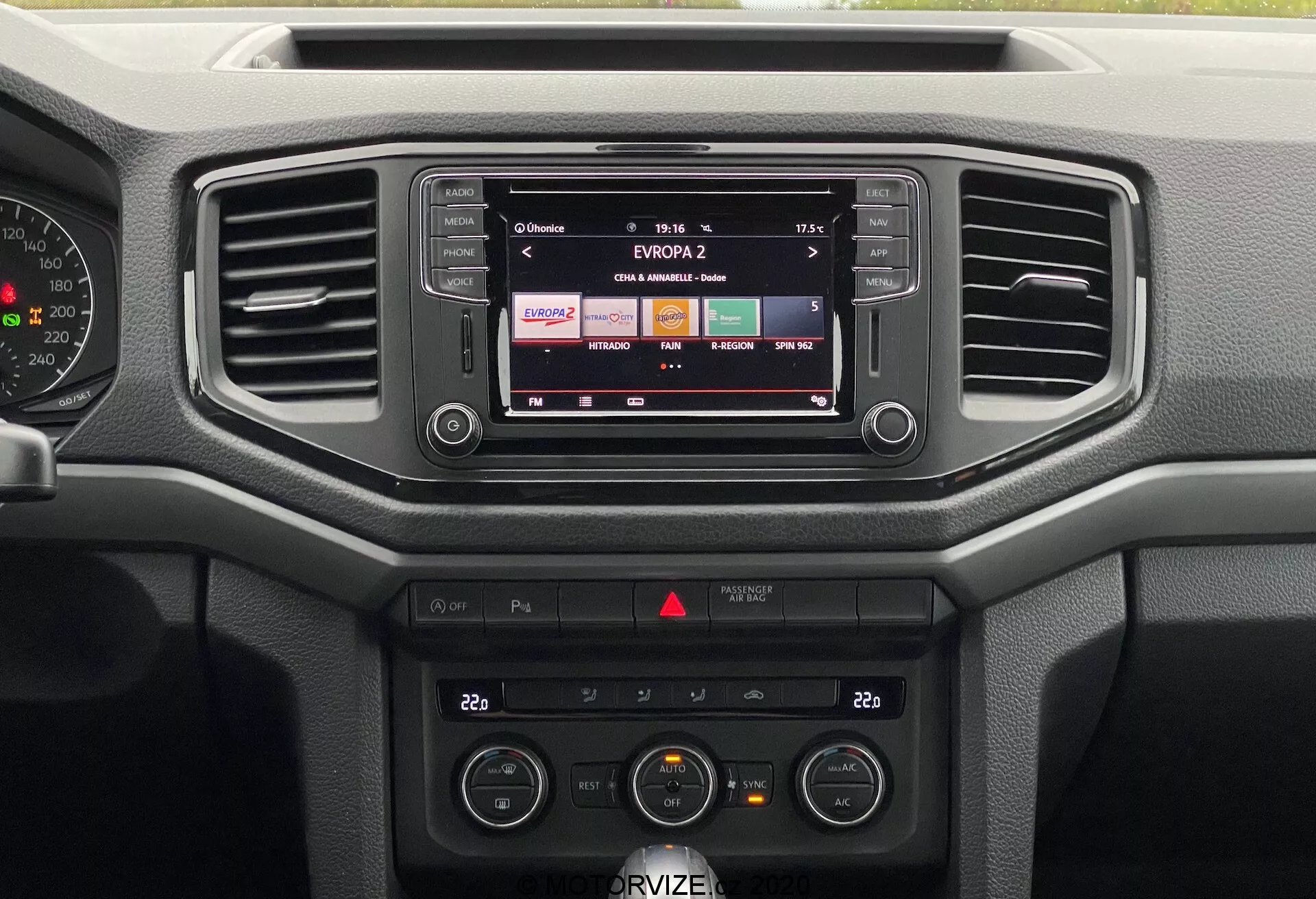 Obraz przedstawia centralną część deski rozdzielczej VW Nutzfahrzeuge Amarok 2016. Z frontowej perspektywy można zobaczyć system infotainment z wyświetlaną stacją radiową, dwa poziome nawiewy i różne przyciski. System audio i sterowanie klimatyzacją, w tym regulacja temperatury i wentylatora, są wyraźnie widoczne. Deska rozdzielcza skupia się na funkcjonalności z łatwymi w użyciu przyciskami i pokrętłami, wykonanymi głównie z twardych tworzyw sztucznych, odpowiednimi do codziennego użytku, a nie luksusowych wykończeń.