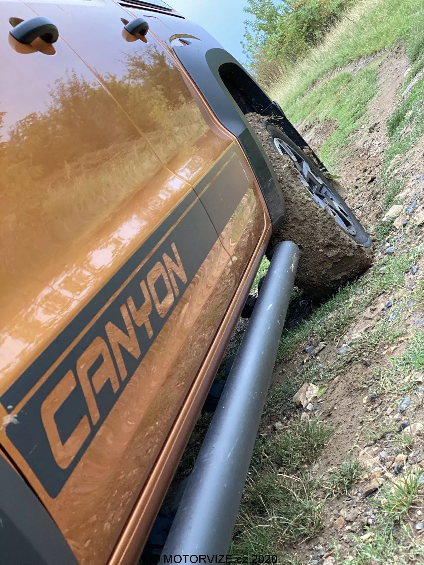 To pochylone pod kątem 45 stopni zdjęcie pokazuje tylną część po stronie pasażera VW-Nutzfahrzeuge Amarok 2016. Skupia się na bliskich szczegółach od tylnej wnęki koła do tylnej części ładunkowej, podkreślając solidne czarne osłony nadkoli i wytrzymałe opony terenowe. Pojazd posiada grafikę z napiszem 'CANYON' i znajduje się obok polnej drogi z kołem zamontowanym na dużym kamieniu, co pokazuje jego terenowe możliwości. Odbijający się metaliczny brązowy lub miedziany kolor nadwozia jest wyraźny, łapiąc odblaski światła. Znacznik czasu 'MOTORVIZE.cz 2020' sugeruje, że zdjęcie zostało zrobione do publikacji motoryzacyjnej.