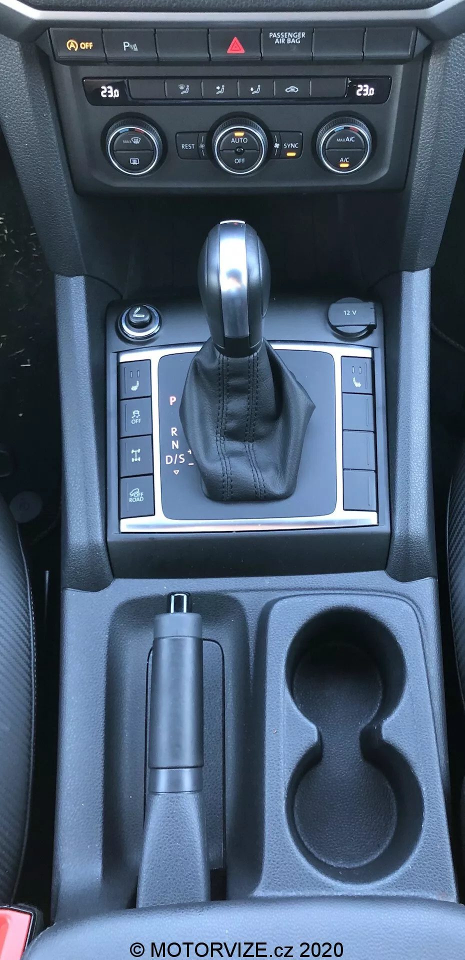 Ten obraz przedstawia konsolę centralną vw-nutzfahrzeuge amarok 2016, wykonany z perspektywy z góry, mniej więcej z punktu widzenia kierowcy lub pasażera z przodu. Fotografia uchwyca wnętrze samochodu, w szczególności dźwignię zmiany biegów, kontrolki konsoli centralnej i schowki. Na zdjęciu znajdują się pokrętła i przyciski systemu klimatyzacji, dźwignia zmiany biegów pokryta skórą osadzona w srebrnym panelu wskazującym skrzynię automatyczną, kontrola elektronicznego hamulca postojowego, schowek, dwa uchwyty na kubki oraz dodatkowy schowek w kierunku tylnych siedzeń. Widoczne materiały to plastik, metaliczne akcenty i skóra, odzwierciedlając połączenie funkcjonalności i estetyki.