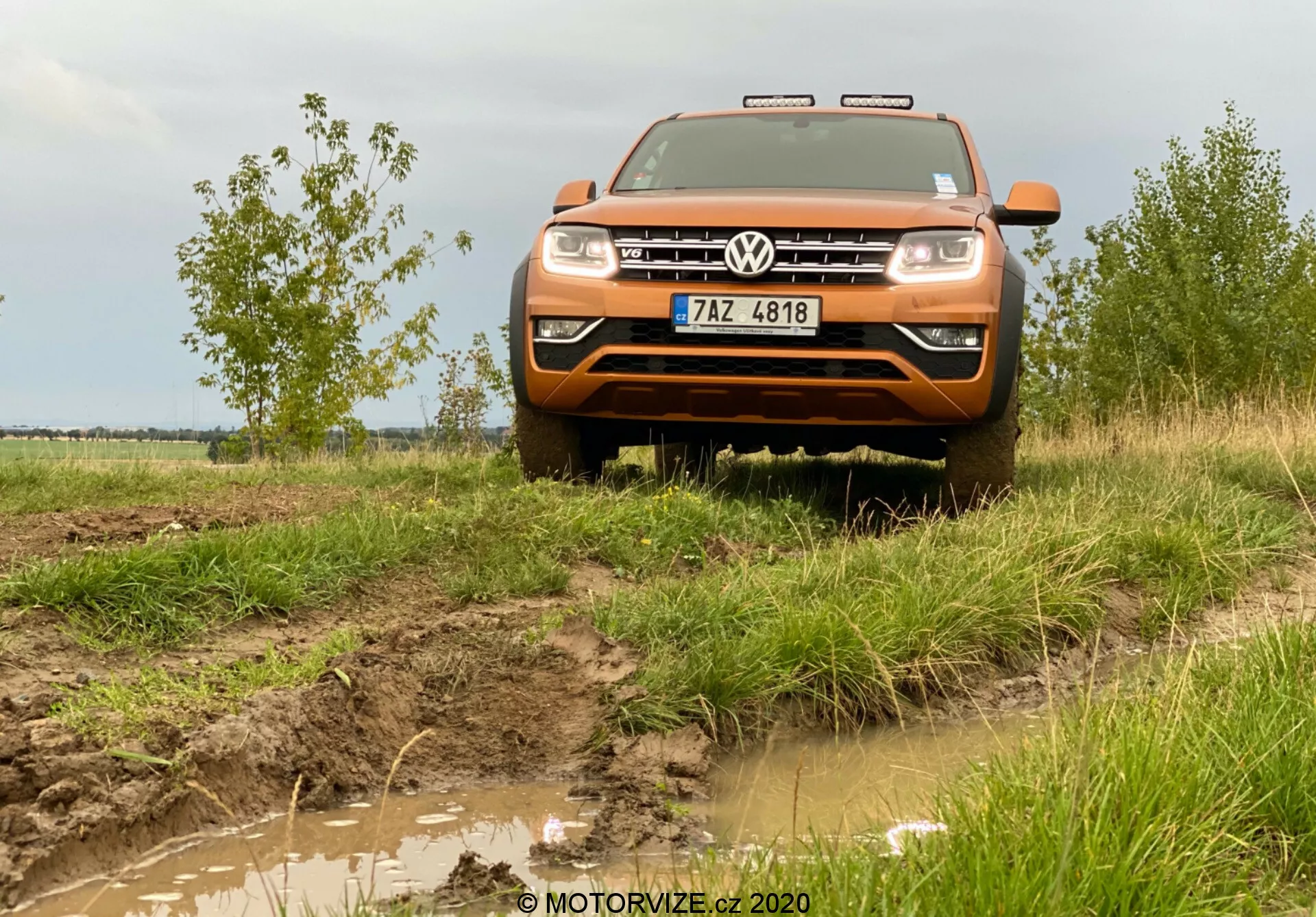 Widok z przodu pod niskim kątem na pomarańczowego Volkswagena Amaroka 2016, średniej wielkości pickupa, znanego z terenowych możliwości. Zdjęcie przedstawia przód pojazdu, w tym maskę, grill, zderzak, część błotników, reflektory oraz logo Volkswagena wyeksponowane na środku grille. Nad grillem widoczne są chromowane akcenty lub LEDowe światła dzienne. Błotnisty teren i trawa wkoło podkreślają terenowy charakter pojazdu, z dodatkowymi światłami na dachu, przednią płytą ochronną i wiejskim tłem z zachmurzonym niebem dopełniającym przygodowy klimat. Obraz jest zrobiony z odległości około dwóch do trzech metrów, dając szczegółowy widok na przednie cechy pojazdu i jego interakcję z otoczeniem.