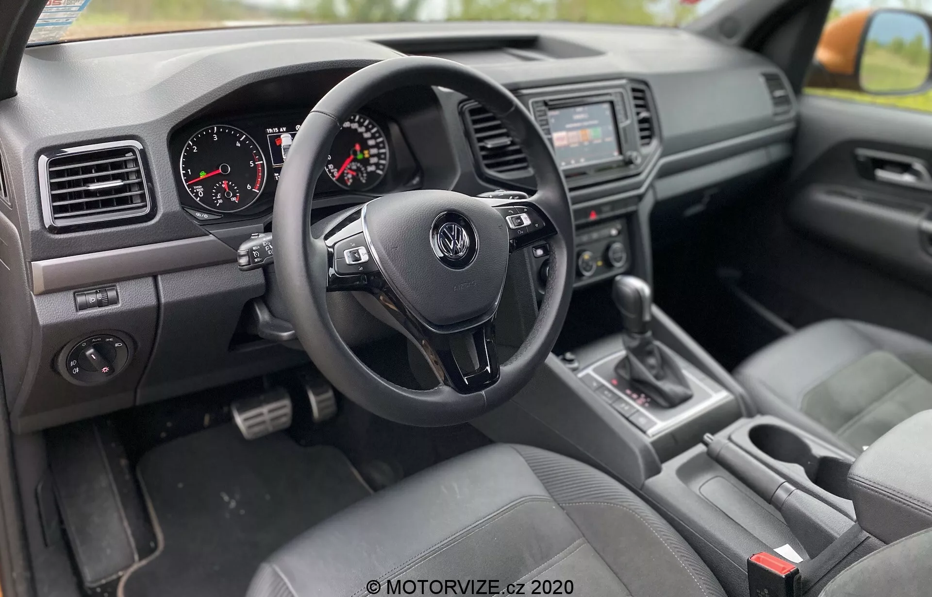 Widok wnętrza VW Nutzfahrzeuge Amarok 2016 z perspektywy pasażera, z prawostronnym układem kierowniczym z trójramienną kierownicą Volkswagen z przyciskami sterującymi, zestawem wskaźników z analogowym prędkościomierzem i obrotomierzem, konsolą środkową z systemem audio i ekranem multimedialnym, dźwignią zmiany biegów w pozycji 'P', siedzeniami tapicerowanymi tkaniną, deską rozdzielczą z otworami wentylacyjnymi i plastikami z fakturą oraz panelem drzwi kierowcy z przyciskami sterowania szybami i regulacją lusterek.