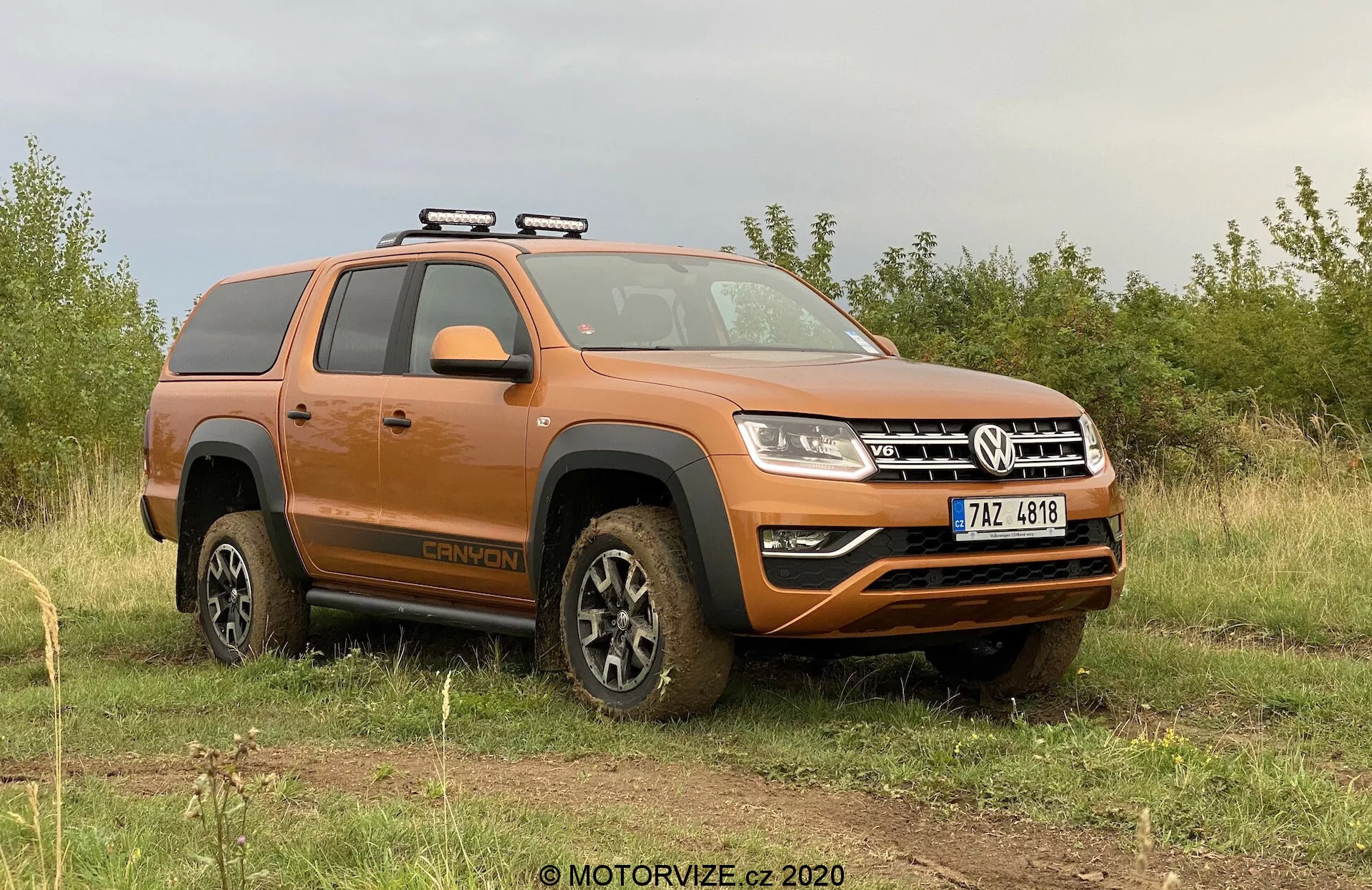 Obraz przedstawia pomarańczowy pickup Volkswagen Amarok 2016, zaparkowany na nierównym trawiastym terenie z chmurami na niebie w tle. Perspektywa z boku-przodu skupia się na stronie kierowcy, pokazując profil pojazdu i część przodu. Widoczne kluczowe cechy to bagażnik dachowy, koła pokryte błotem sugerujące niedawną aktywność terenową, stopnie boczne, konfiguracja z podwójną kabiną, nowoczesny design reflektorów, a także przedni zderzak z światłami przeciwmgielnymi i dolną płytą ochronną. Pojazd emanuje surowym wyglądem użytkowym terenowym z nowoczesnymi elementami technologicznymi.