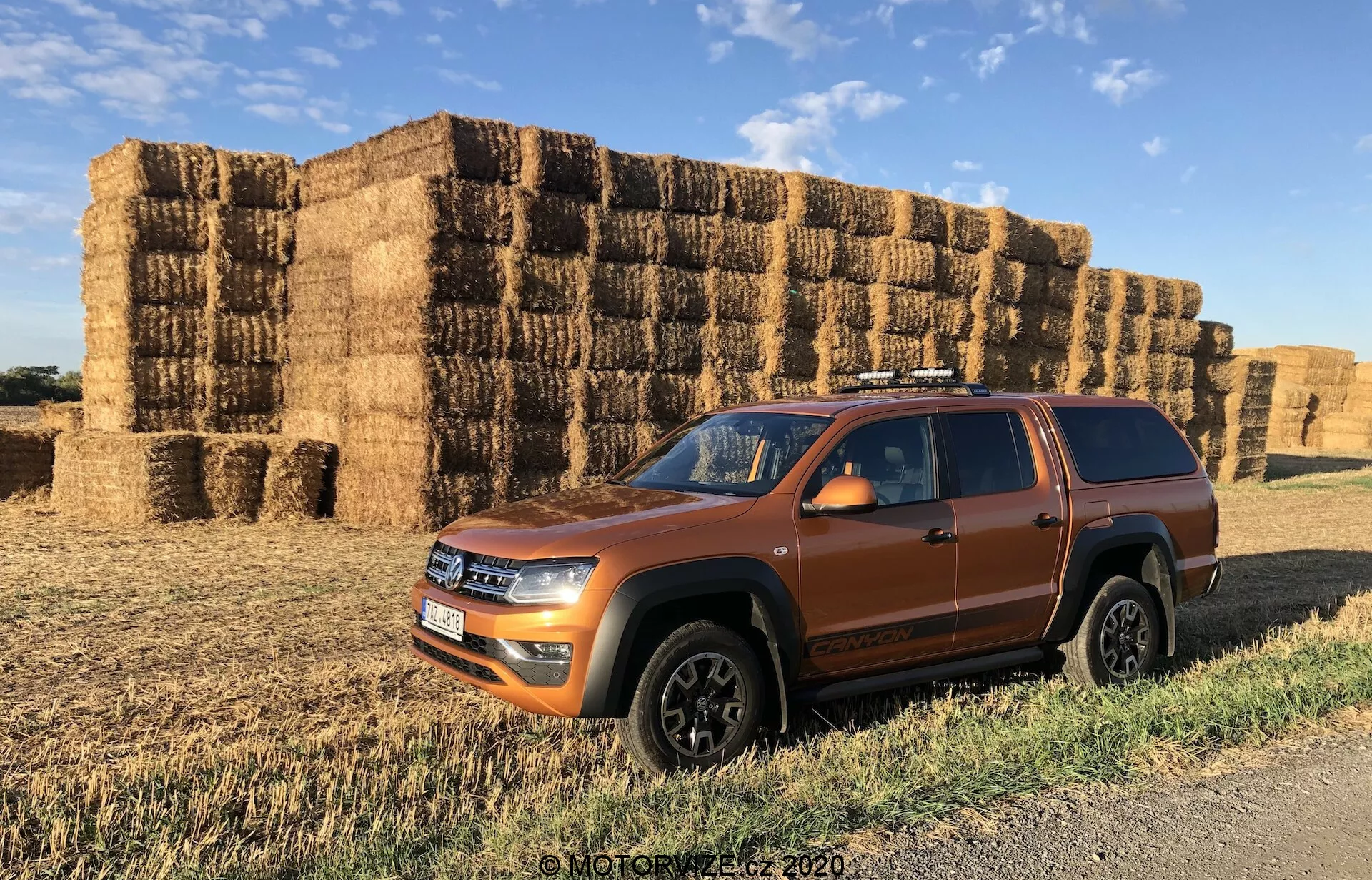 Widok z profilu bocznego pomarańczowego SUV-a VW Nutzfahrzeuge Amarok 2016 podkreślający przednią fasadę, stronę kierowcy i dach. Opływowy kształt nadwozia, reflektory LED, solidne nadkola i czarne wykończenie kół sugerują nowoczesny, sportowy design. Relingi dachowe i widoczny prześwit wzmacniają jego wielofunkcyjność i zdolności terenowe. Umieszczony na gruntowej drodze z belami siana w tle, obraz podkreśla solidne, wiejskie otoczenie pojazdu.