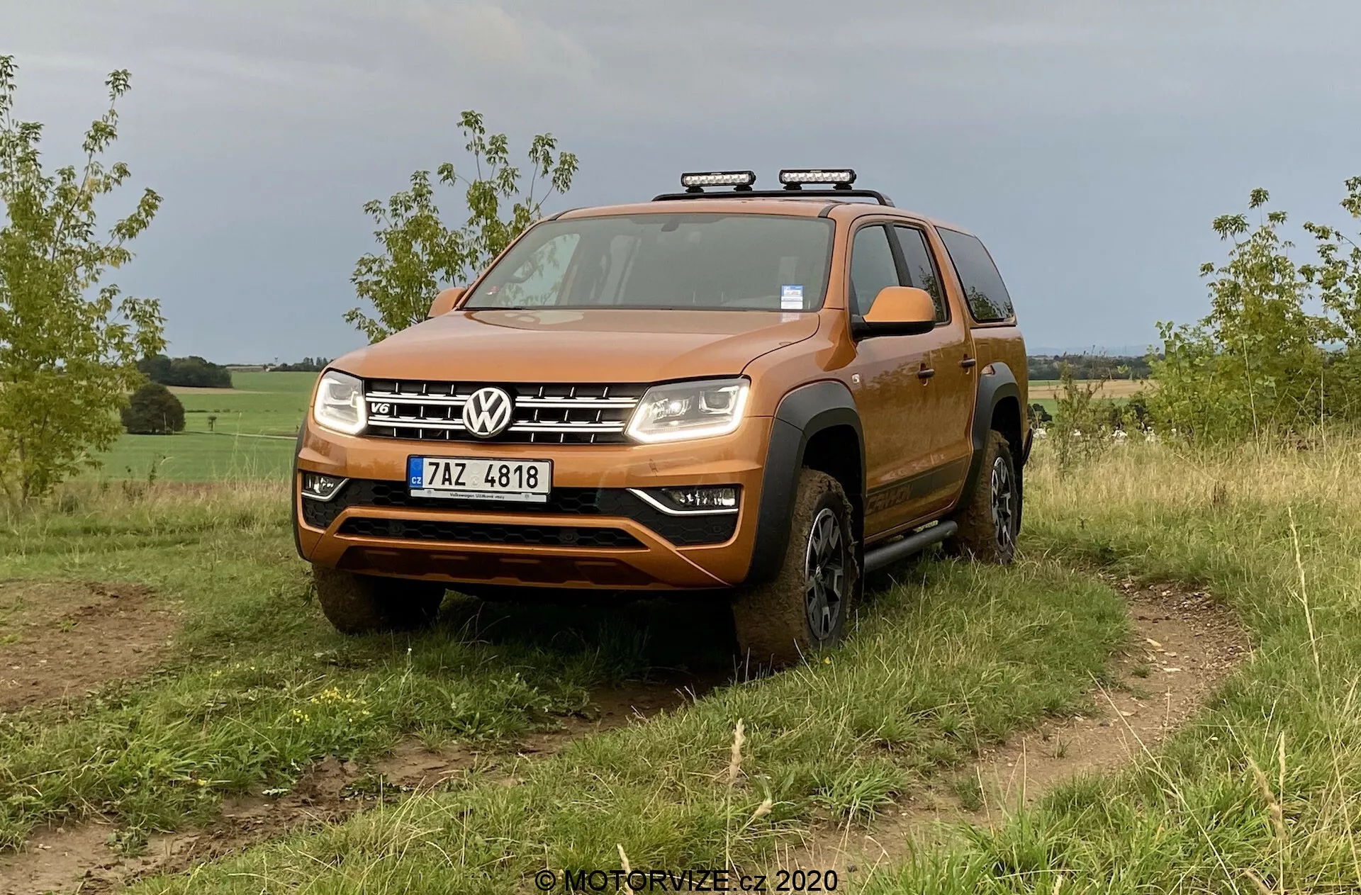 Pomarańczowy SUV Volkswagen, konkretnie model VW Amarok 2016, jest pokazywany na trawiastym terenie off-road. Zdjęcie z niskiego do średniego kąta z przodu podkreśla jego wytrzymały design, odpowiedni do jazdy terenowej. Pojazd ma wyraźny przedni grill z dwoma chromowanymi prętami, które obramowują centralne logo Volkswagen, znaczny zderzak z możliwą płytą poślizgową, wyraźne kompozytowe reflektory z potencjalnymi żarówkami LED lub halogenowymi oraz rozszerzone nadkola mieszczące opony terenowe. Na dachu zamontowane są dodatkowe światła, co sugeruje zwiększoną funkcjonalność do jazdy nocą. Widoczne szczegóły obejmują przednią szybę, relingi dachowe, lusterka boczne z wbudowanymi kierunkowskazami i przednią tablicę rejestracyjną. Pojazd wydaje się czysty z pewnymi zabrudzeniami na dolnych częściach i kołach, co pasuje do użytkowania w terenie. Tło sugeruje wiejskie lub pół-wiejskie otoczenie.