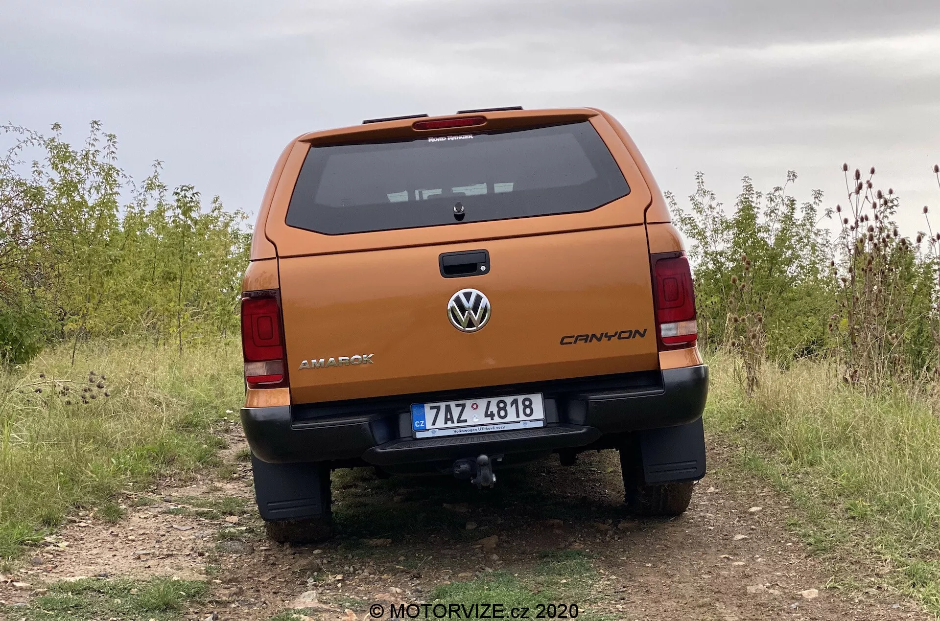 Profesjonalny widok tylny na pickup Volkswagen Amarok 2016 z niskiego kąta, podkreślający solidną klapę tylną z oznaczeniem 'AMAROK', logo Volkswagena pośrodku, wskaźnik poziomu wyposażenia 'CANYON', tablicę rejestracyjną, szczegółowe światła tylne, zderzak z czujnikami parkowania i stopniem, zaczep holowniczy, zaparkowany na nieutwardzonej drodze, wskazujący na możliwości terenowe.