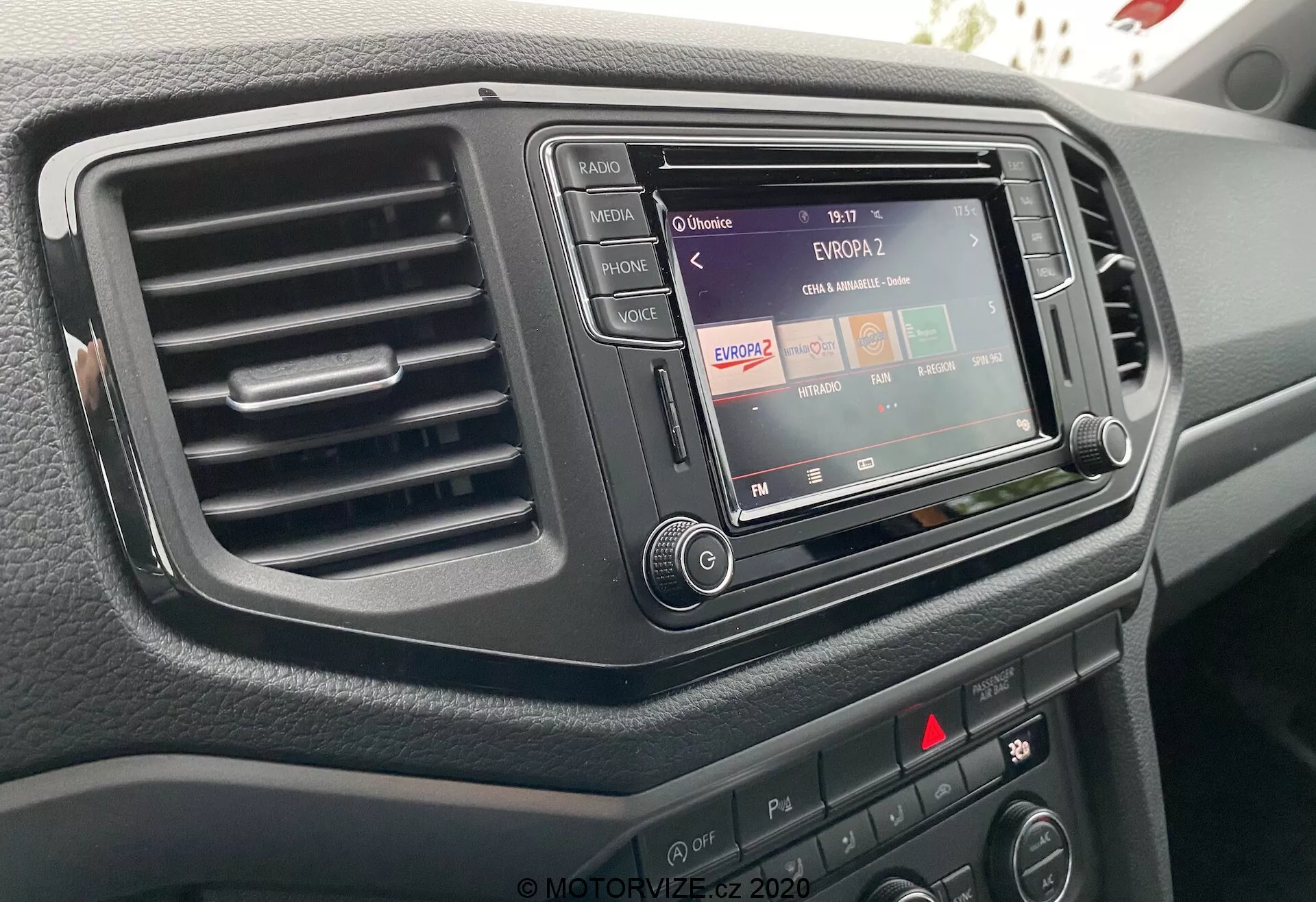 Zbliżenie konsoli centralnej VW Nutzfahrzeuge Amarok 2016 z perspektywy kierowcy, ukazujące system infotainment z funkcją radia, pojedynczy otwór wentylacyjny po lewej stronie oraz różne przyciski i pokrętła sterujące. System infotainment jest osadzony w ciemnym, eleganckim wykończeniu, a powierzchnia deski rozdzielczej ma fakturę.