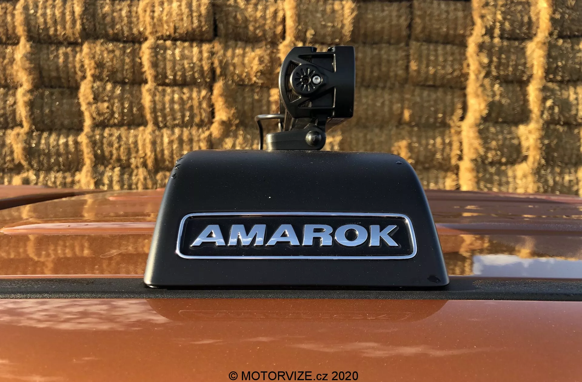 Zbliżenie zdjęcia pickupa Volkswagen Amarok 2016 z widoczną kamerą cofania zamontowaną na wysoko umieszczonym światle hamowania z napisem 'AMAROK' w srebrnych wypukłych literach na metalicznie brązowym nadwoziu, na zewnątrz na tle wzoru z cegieł.