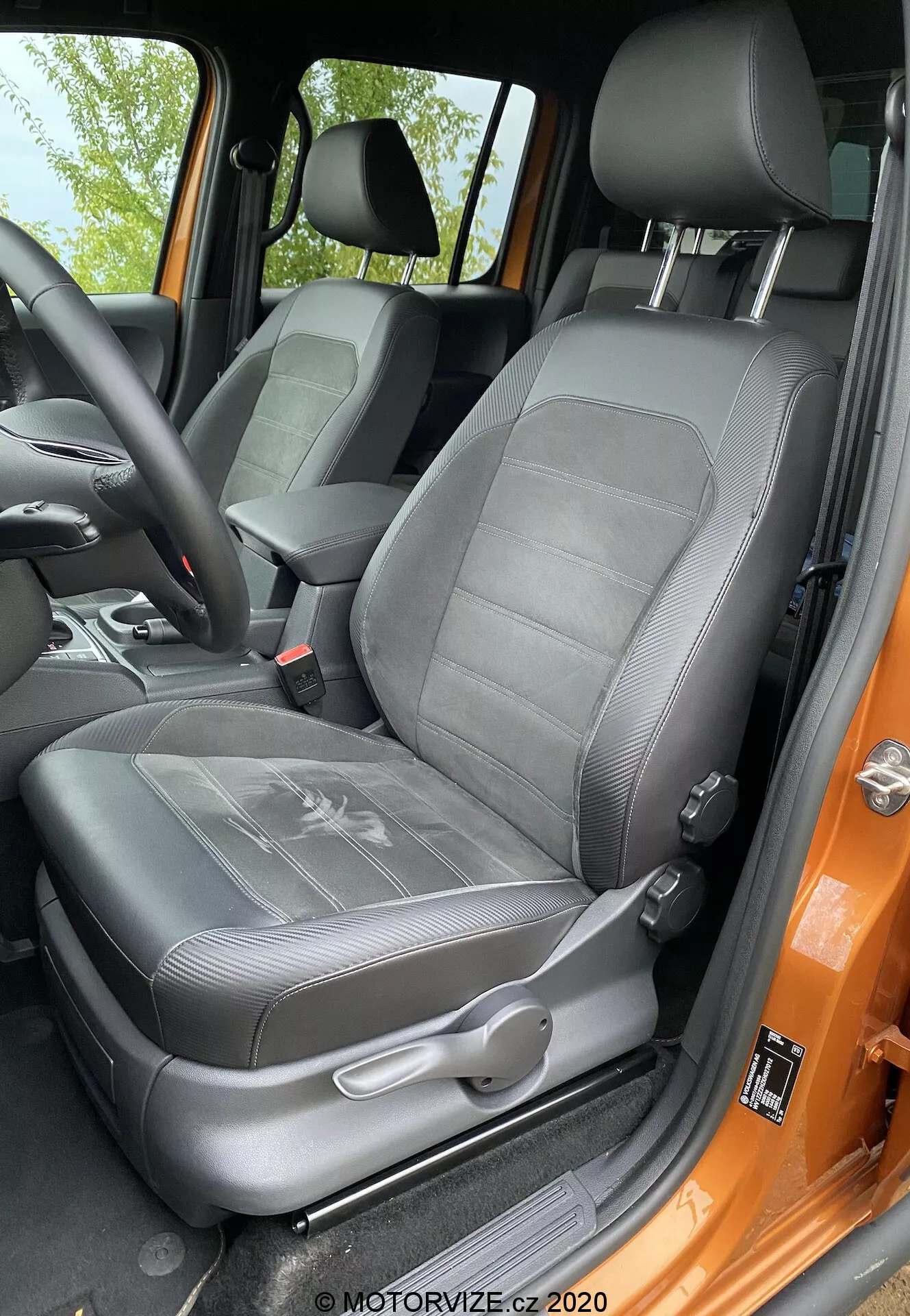 Zbliżenie na fotel kierowcy VW-nutzfahrzeuge Amarok 2016 z perspektywy osoby stojącej na zewnątrz otwartych drzwi. Czarny tapicerowany skórą fotel ma rowkowany wzór i zintegrowany regulowany zagłówek. Widoczne są ręczne dźwignie regulacyjne oraz mechanizm trzypunktowego pasa bezpieczeństwa. Boczne wsparcie wskazuje na boczne podparcie, a pomarańczowa rama drzwi kontrastuje z czarnym wnętrzem. Fotel wydaje się zadbany i jest oświetlony naturalnym światłem dziennym.