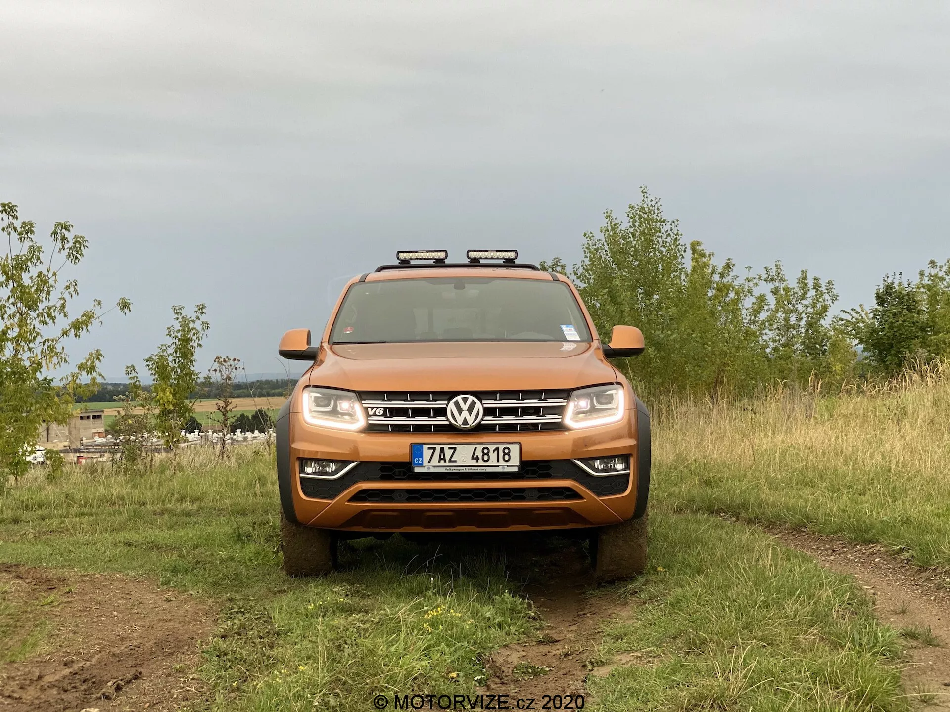 Widok z przodu SUV-a Volkswagen Amarok uchwycony w scenerii plenerowej. Pomarańczowy pojazd ma wyraźne logo Volkswagena na grillu, poziome listwy, standardowe reflektory i dodatkowe światła pomocnicze zamontowane na dachu. Obraz podkreśla możliwości terenowe z częściowo widocznymi, wytrzymałymi oponami i zabrudzeniami na powierzchni pojazdu. Jest umieszczony na naturalnym terenie z rozmytym tłem, oświetlony rozproszonym światłem z zachmurzonego nieba.
