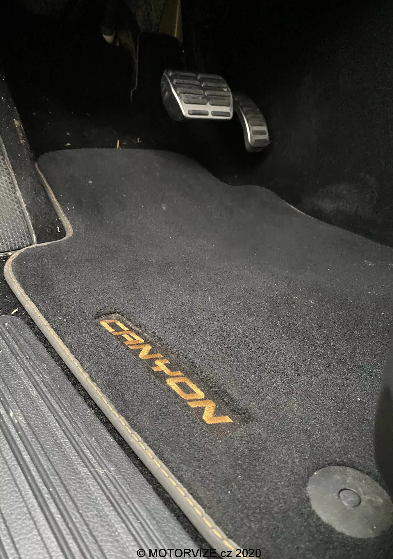 Zbliżenie na obszar podłogi po stronie kierowcy w VW-Nutzfahrzeuge Amarok 2016, pokazujące ciemną matę podłogową z haftem 'ENYON'. Widoczne są metaliczne pedały gazu i hamulca, przy czym pedał hamulca znajduje się wyżej, co sugeruje, że jest zaciągnięty. Mata jest konturowana, aby pasowała do przestrzeni, z hakiem mocującym po prawej stronie. Mata kontrastuje z ciemnym otoczeniem, wskazując na ciemne wnętrze. Scena jest słabo oświetlona, prawdopodobnie zrobiona w nocy lub w warunkach słabego oświetlenia.