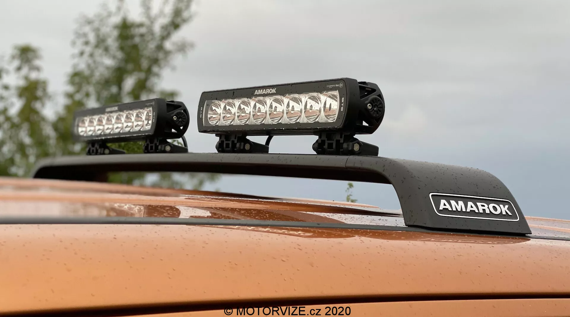 Zbliżenie skierowane ku górze pokazujące dach Volkswagena Amaroka 2016 z zamontowanym na dachu oświetleniem, dwoma wysokowydajnymi listwami LED na wsporniku z logo 'AMAROK', metaliczne pomarańczowe wykończenie; idealne do cech pojazdów terenowych lub akcesoriów pojazdów.