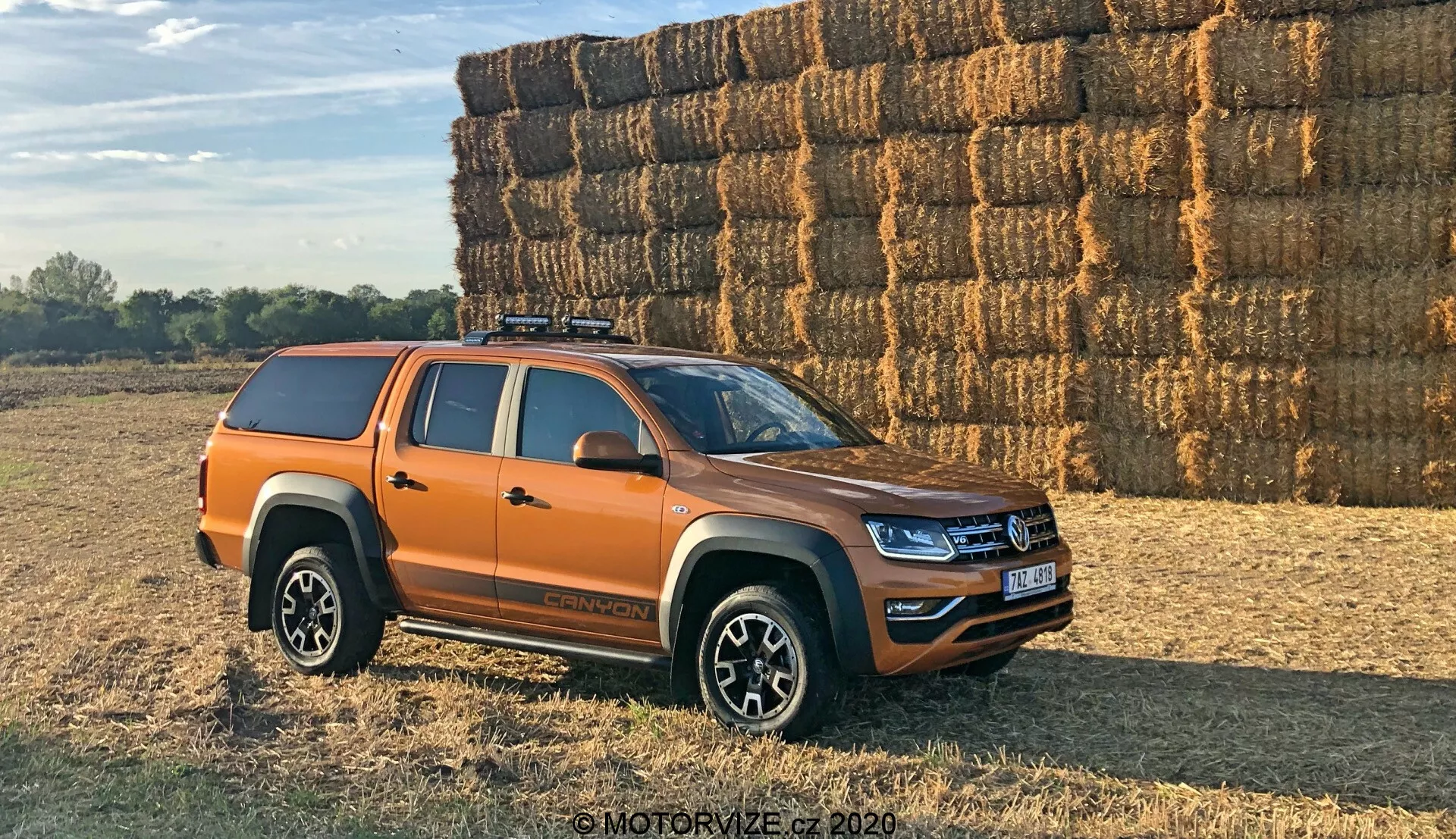 Widok z boku pomarańczowego pickupa Volkswagen Amarok zaparkowanego na zewnątrz na nierównej, prawdopodobnie ziemnej lub trawiastej powierzchni. Pojazd jest umieszczony pod kątem 30 stopni w stosunku do kamery, pokazując profil boczny oraz częściowy widok na przednią kratkę i reflektory. Nowoczesny pickup średniej wielkości ma świeże linie projektowe oraz oświetlenie LED, sugerujące niedawny model. Widać cztery drzwi, co wskazuje na konfigurację z podwójną kabiną, wieloramienne felgi aluminiowe, opony terenowe, twardą osłonę na skrzynię ładunkową oraz system bagażnika dachowego. Lusterka boczne prawdopodobnie zawierają kierunkowskazy. Zdjęcie wykonane w ciepłym, dziennym świetle z długimi cieniami, dużą stertą bel słomy w tle wskazuje na wiejskie otoczenie.