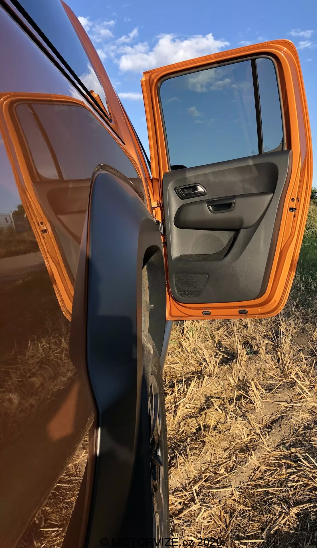 Zbliżenie na częściowo otwarte przednie drzwi pasażera pomarańczowego VW Nutzfahrzeuge Amarok 2016, ukazujące jasnopomarańczową farbę zewnętrzną i kontrastujący czarny wewnętrzny panel drzwi z wbudowanymi elementami sterującymi. Samochód jest zaparkowany ukośnie, prezentując boczny profil z drzwiami otwartymi pod kątem 70 stopni, podkreślając lusterko boczne z czarną obudową. Odbicia na drzwiach pokazują czyste niebo i płaski teren rolniczy, co podkreśla wiejskie otoczenie.