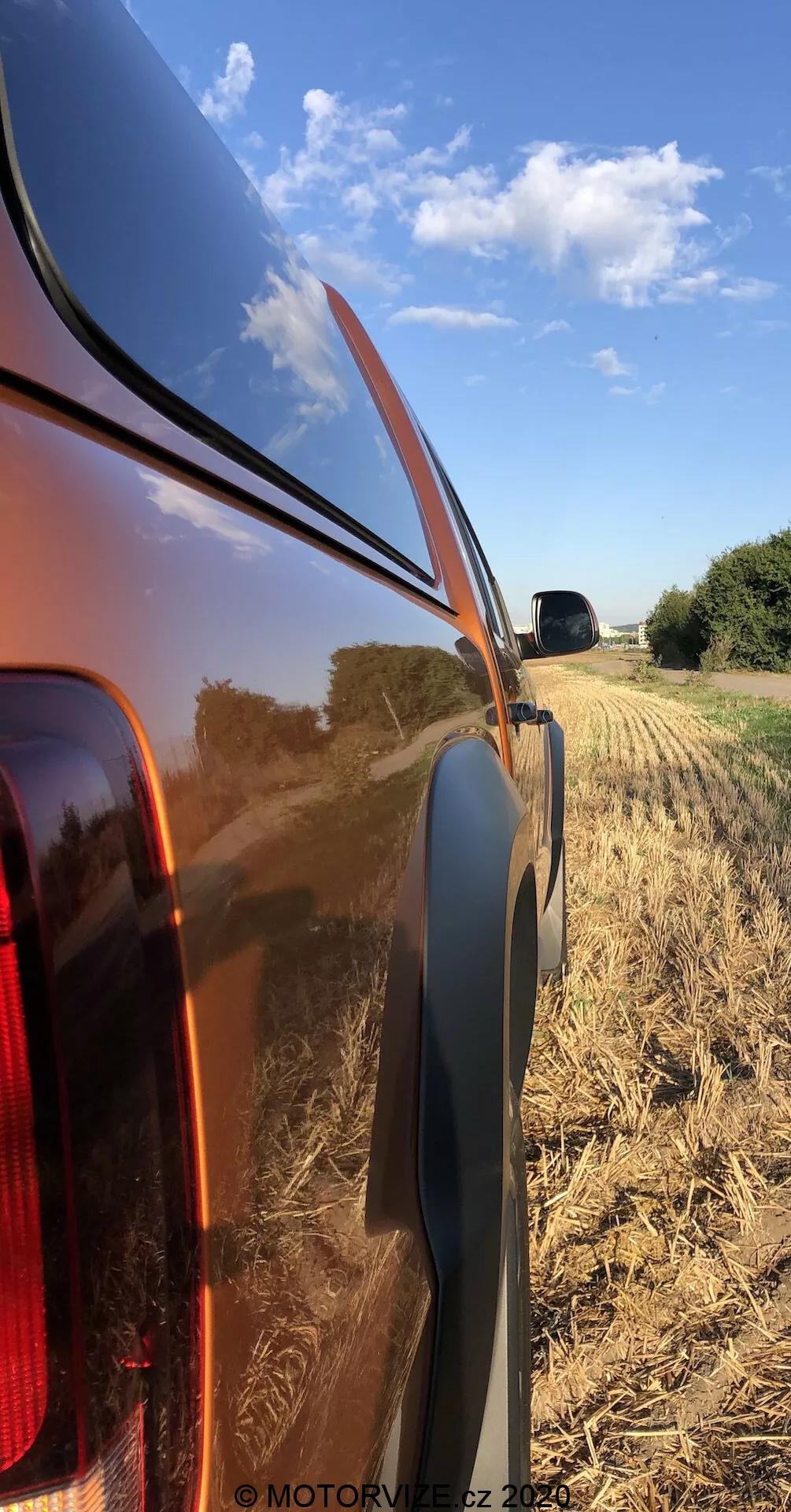 Zbliżenie na prawy tylny panel 2016 VW Nutzfahrzeuge Amarok, podkreślający błyszczące, odblaskowe metalowe nadwozie i zintegrowane tylne światło z przezroczystą soczewką. Zdjęcie, wykonane pod niskim kątem, ukazuje linię pasa, czarną plastikową listwę i tło wiejskiego żniwnego pola z jasnym niebem.