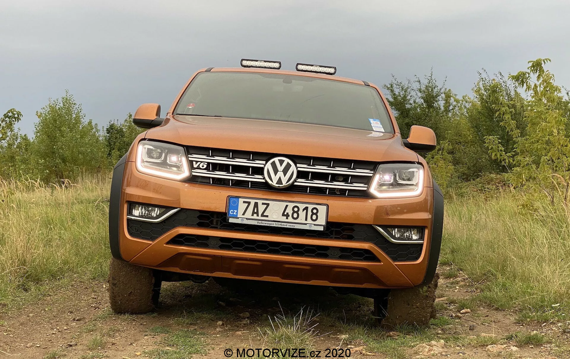 Widok z przodu metalicznego pomarańczowego pickupa Volkswagen Amarok, ukazującego swoją solidną postawę i zdolności off-roadowe z wyróżniającym się grillem z emblematem VW i odznaką V6. Zdjęcie, zrobione z niskiego kąta, ukazuje pełną szerokość przedniej części pojazdu, w tym reflektory i dodatkowe światła terenowe, z naturalnym tłem pola i drzew podkreślającym solidność pickupa. Widoczne funkcje obejmują solidny przedni zderzak, płytę ochronną, aktywowany zaawansowany system oświetlenia i tablicę rejestracyjną Republiki Czeskiej.