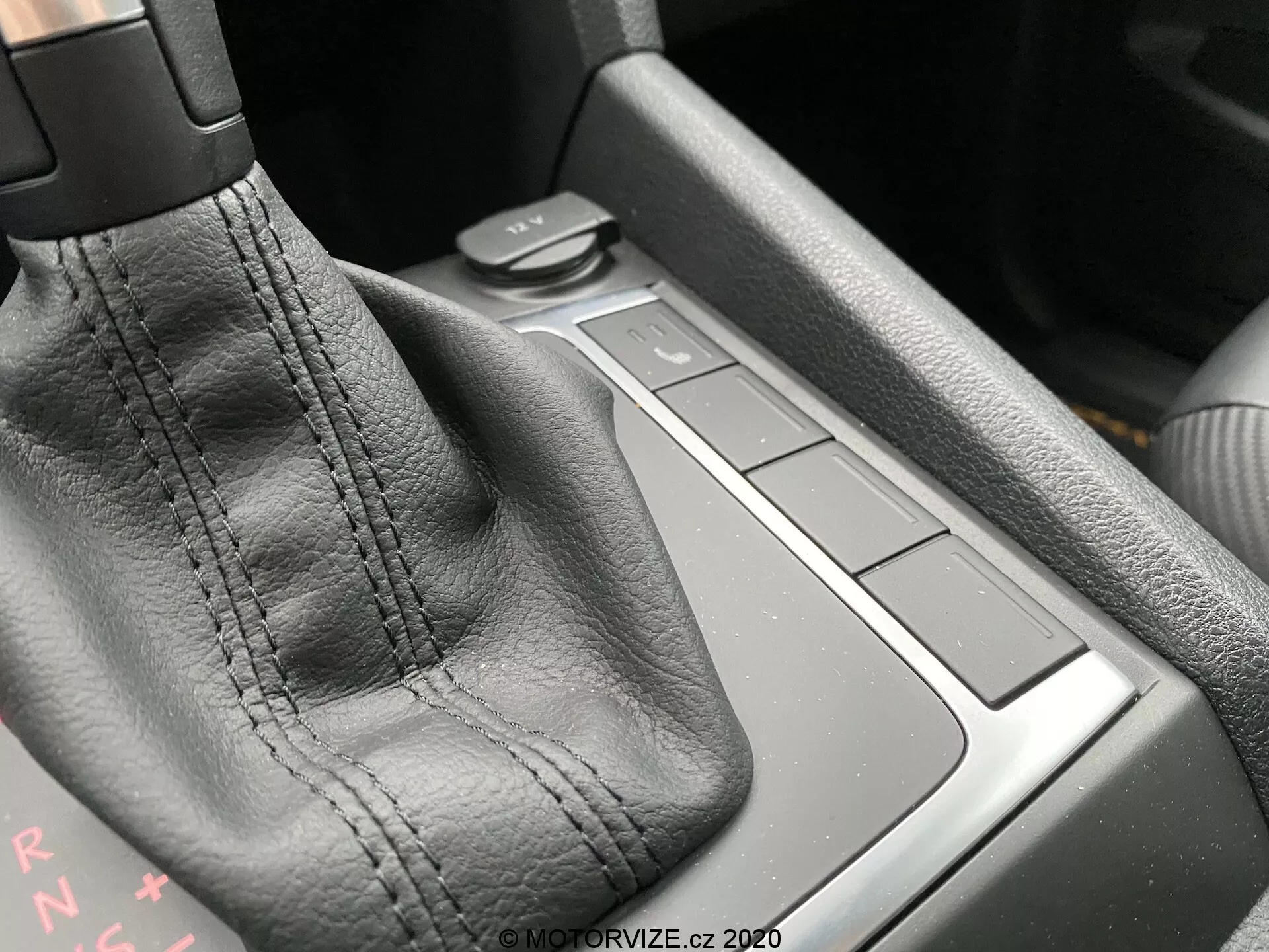 Zbliżenie konsoli środkowej VW Nutzfahrzeuge Amarok 2016, koncentrujące się na obszarze dźwigni zmiany biegów. Głównym elementem jest dźwignia zmiany biegów pokryta czarną skórą z widocznymi szwami. Litery 'PRNDS' za dźwignią wskazują na automatyczną skrzynię biegów. Po prawej znajdują się cztery kwadratowe przyciski na równi z powierzchnią konsoli, prawdopodobnie do sterowania różnymi funkcjami samochodu. Obszar za przyciskami może być małym schowkiem. Materiały i jakość wykonania sugerują wysoki poziom rzemiosła. Światło otoczenia z lewego górnego rogu podkreśla detale.