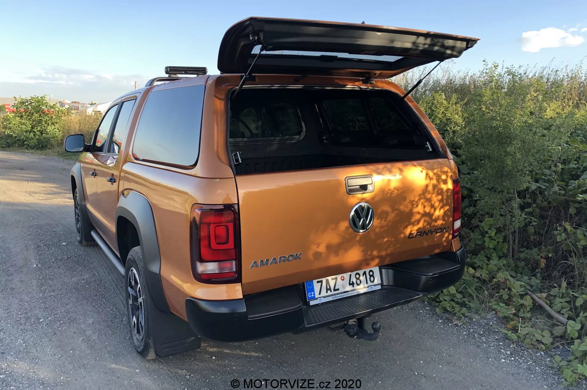 Tylny trzykątny widok Volkswagena Amarok 2016 w metalicznym pomarańczowym kolorze, podkreślający otwartą osłonę bagażnika, hak holowniczy, drzwi drugiego rzędu podwójnej kabiny i część tylnego prawego koła i zderzaka, umiejscowiony na wiejskim terenie na zmierzchu. Zdjęcie od MOTORVIZE.cz, 2020.