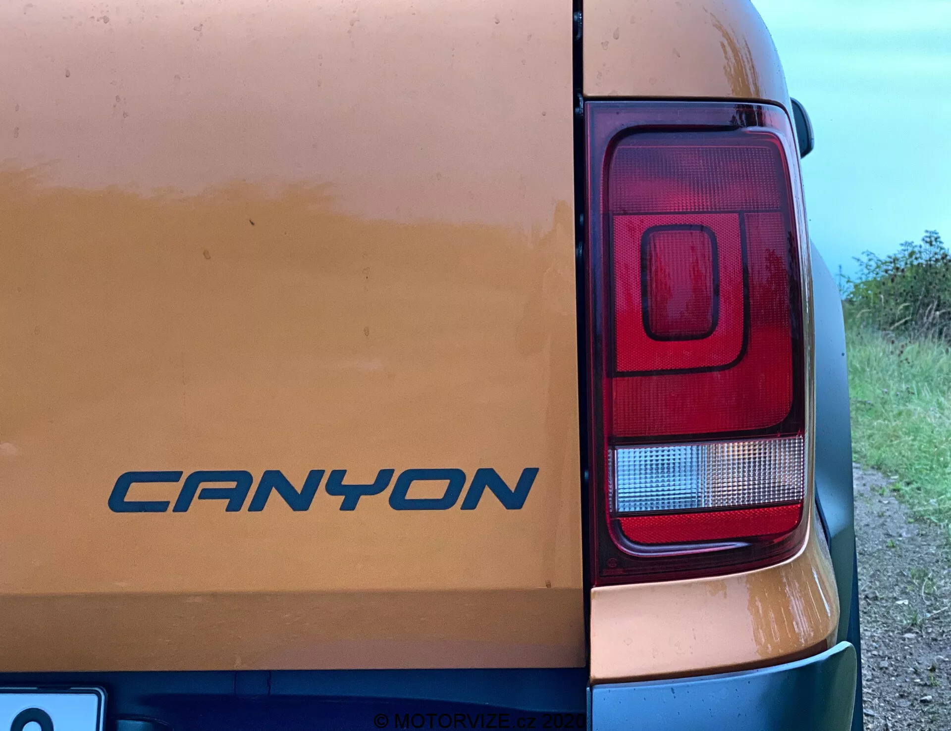 Zbliżenie na prawy tylny róg pojazdu VW Nutzfahrzeuge Amarok 2016. Na tylnych drzwiach widoczny jest emblemat 'CANYON' pośród śladów brudu, co sugeruje użytkowanie na nieutwardzonych drogach. Fotografia wyraźnie podkreśla prostokątny nowoczesny klaster świateł tylnych z sekcjami dla świateł hamowania, cofania i prawdopodobnie kierunkowskazów. Zdjęcie wykonane pod kątem bocznym około 45 stopni ukazuje częściowo karoserię pojazdu z wiejskim lub półwiejskim tłem.