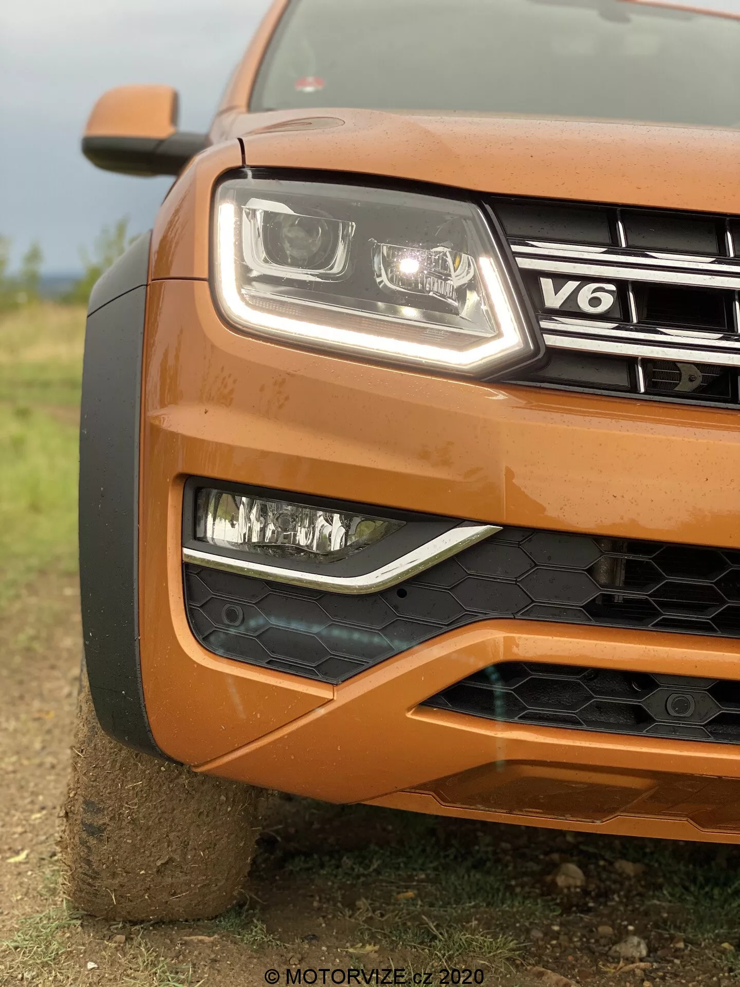 Zbliżenie na lewy przedni róg pomarańczowego VW Nutzfahrzeuge Amarok z 2016 roku, ukazujące reflektor z nowoczesną, przezroczystą soczewką i technologią LED, światła przeciwmgielne w czarnym wzorzystym okręgu, częściowo widoczny grill z emblematem V6, zderzak przedni z czarną strukturą plastra miodu, konturowany kaptur oraz częściowo widoczne duże koło, z rozmytym tłem podkreślającym teksturę i solidny design samochodu