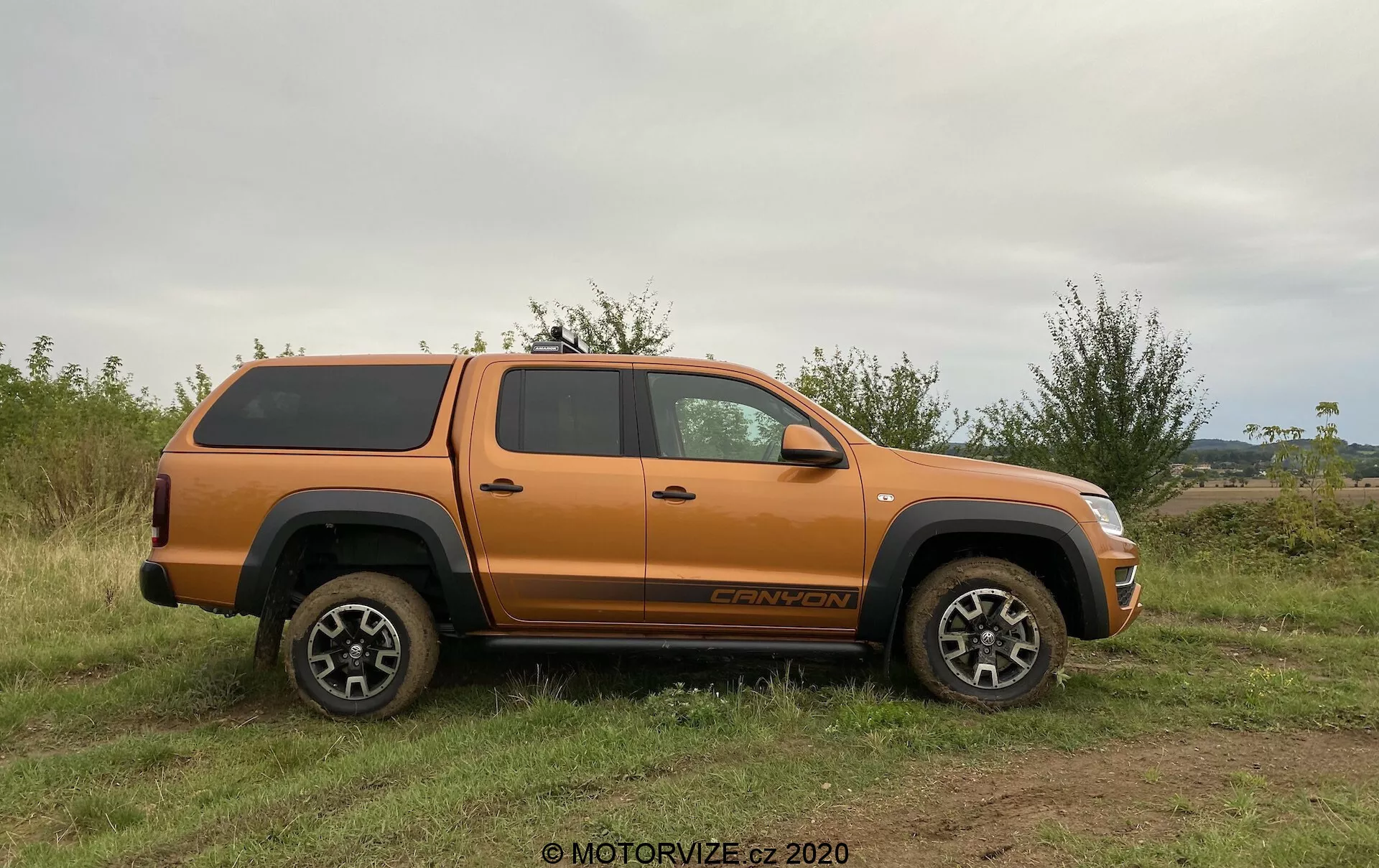 Profil boczny pomarańczowego pickupa VW Nutzfahrzeuge Amarok 2016 z twardą pokrywą na skrzyni, zaparkowanego na nierównym, trawiastym terenie. Pojazd ma duży prześwit, solidne koła i opony oraz wieloszprychowy design z lekkich stopów, przystosowany do jazdy terenowej. Widoczne elementy to lusterka boczne, klamki drzwi, nadkola, stopnie, przestronna kabina na cztery drzwi, odznaka na przednim kole i napis określający wersję wyposażenia nad tylnym kołem. Naturalne światło i wyrównany kąt podkreślają szczegóły pojazdu bez ostrych cieni, uwydatniając jego zdolności terenowe i bezpieczny system przechowywania ładunku.
