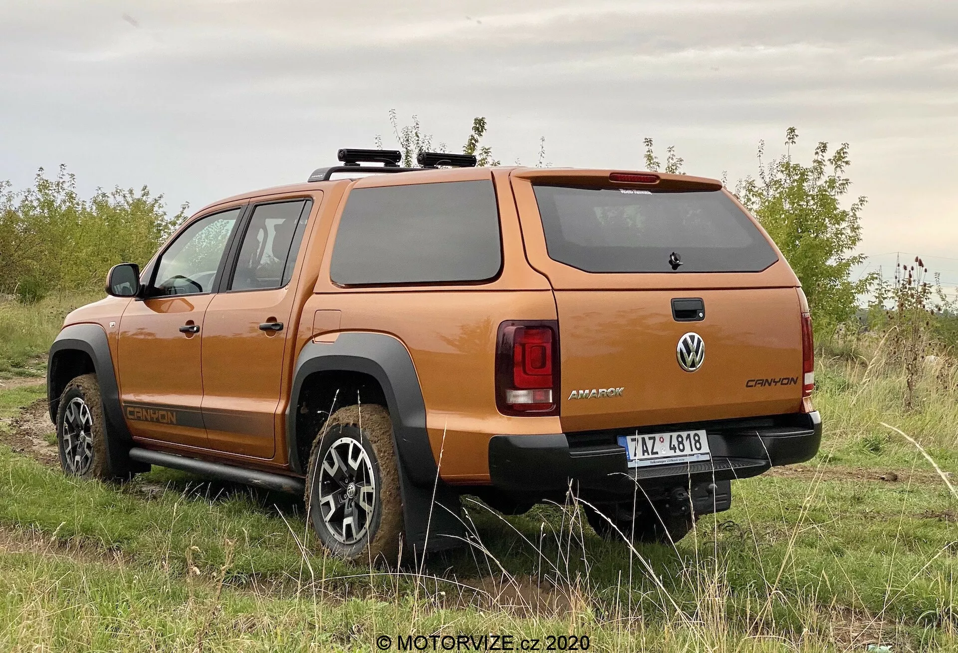 Widok tyłu trzy czwarte pomarańczowego pickupa Volkswagen Amarok 2016, ukazujący design kół, profil boczny, tylne okno, tylne światła, solidny tylny zderzak i sztywną pokrywę przestrzeni ładunkowej, zaparkowanego na nieutwardzonej powierzchni z roślinnością, podkreślający jego terenowe możliwości.