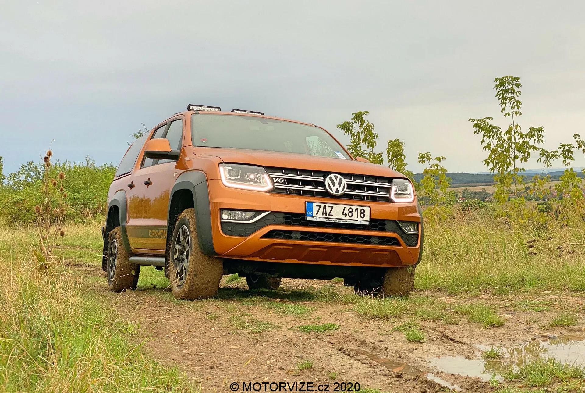 Niski widok na trzy-czwarte przodu Volkswagena Amaroka 2016 na błotnistym polu, prezentujący jego pomarańczowy lakier, przedni grill z logo Volkswagena, reflektory, design kół i zdolności terenowe. Wytrzymałość pojazdu podkreślona jest przez błoto na oponach i dolnych częściach, z przednimi kołami skręconymi w lewo, co uwidacznia wzory bieżnika i wysokość prześwitu. Na miękko oświetlonym tle, o poranku lub późnym popołudniem, obraz podkreśla terenową użyteczność Amaroka i zaawansowany system oświetlenia. Tablica rejestracyjna jest widoczna i powinna być zasłonięta ze względów prywatności.