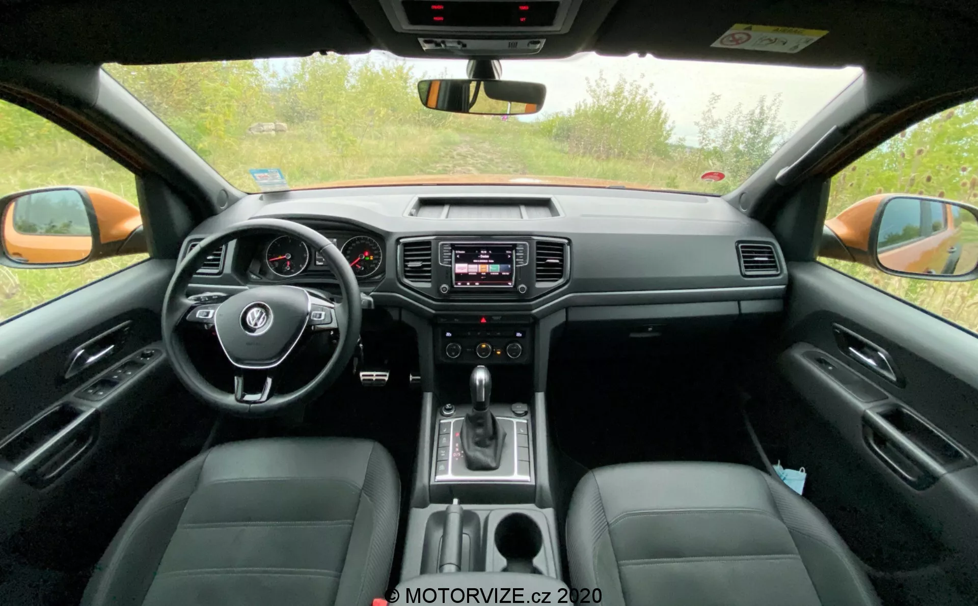 Widok wnętrza przedziału pasażerskiego VW-Nutzfahrzeuge Amarok 2016. Zdjęcie zrobione z perspektywy tylnego siedzenia pokazuje centralny kierownicą z zamontowanymi kontrolkami, analogowy panel instrumentów z małym wyświetlaczem cyfrowym, system infotainment na środku deski rozdzielczej z kontrolkami audio i klimatyzacji, dźwignię zmiany biegów wskazującą na automatyczną skrzynię biegów i skórzane siedzenia. Obraz również uchwyca lusterko wsteczne, zewnętrzne lusterka boczne i przednie okna boczne, podkreślając zarówno technologiczne, jak i wygodne funkcje.