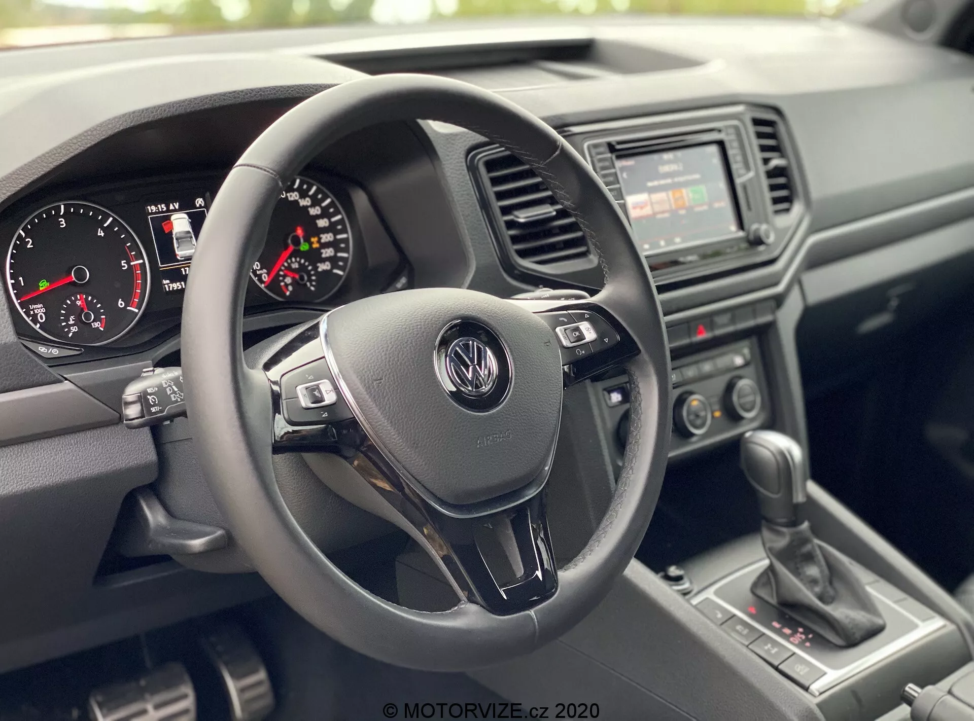 Widok wnętrza VW Nutzfahrzeuge Amarok 2016 z perspektywy pasażera z przodu, pokazujący obszar kokpitu kierowcy. Centralny punkt skupienia to skórzany trójramienny kierownica z logo Volkswagena i wielofunkcyjnymi przyciskami sterującymi, zestaw wskaźników z analogowym prędkościomierzem, obrotomierzem i małym wyświetlaczem cyfrowym, podstawowe radio z ręcznymi przełącznikami klimatyzacji na konsoli środkowej, lewarek manualnej skrzyni biegów oraz materiały z tworzywa sztucznego do deski rozdzielczej i drzwi, podkreślające trwałe i praktyczne wzornictwo.