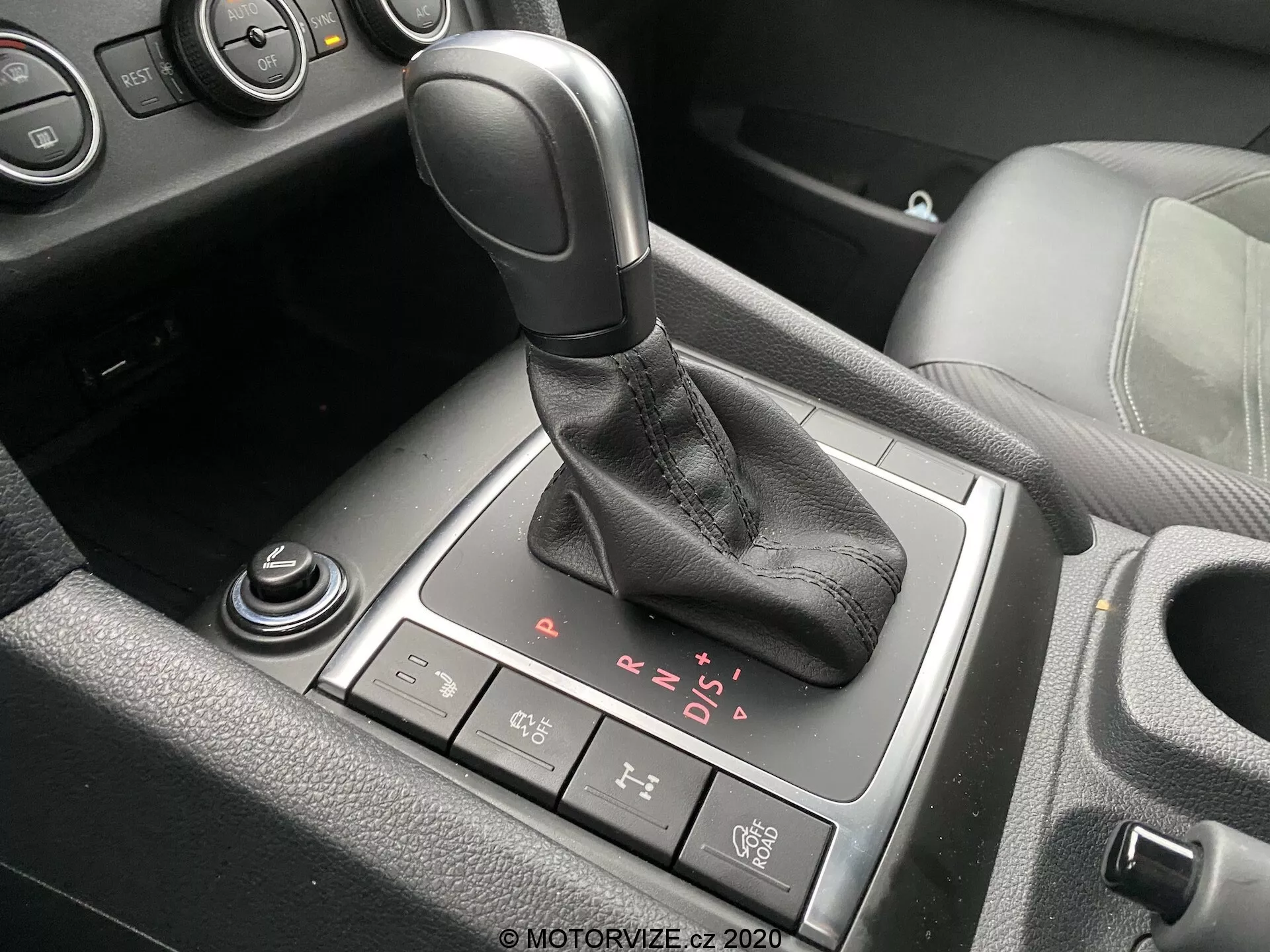 Szczegółowy widok obszaru lewarka zmiany biegów we wnętrzu VW Nutzfahrzeuge Amarok 2016. Automatyczna dźwignia zmiany biegów z czarnym uchwytem i skórzanym mankietem znajduje się centralnie na obrazku, z widocznymi opcjami P, R, N, D oraz trybem ręcznym sekwencyjnym. Za dźwignią zmiany biegów znajduje się przycisk elektronicznego hamulca postojowego, podczas gdy częściowo widoczne pokrętło i inne pokrętło znajdują się odpowiednio z przodu i w lewym dolnym rogu. Widoczne są także fragmentaryczne widoki uchwytu na kubki oraz konsoli środkowej z czarnymi plastikowymi akcentami i możliwymi aluminiowymi lub chromowanymi wykończeniami, co wskazuje na nowoczesny pojazd z wnętrzem średniej lub wyższej klasy.