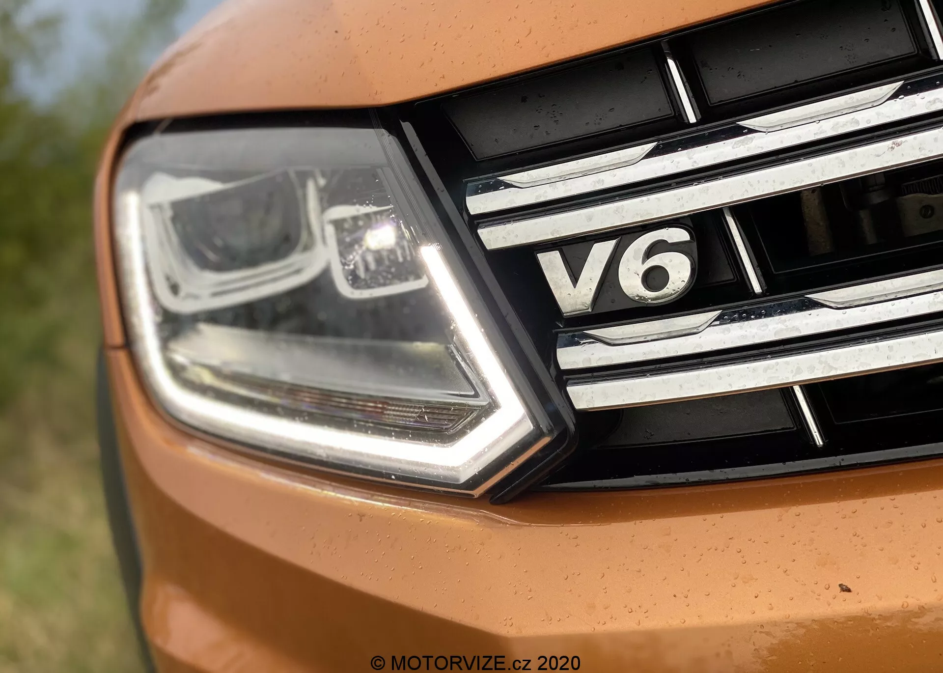 Zbliżenie lewej przedniej części VW Nutzfahrzeuge Amarok 2016. Obraz uchwytuje nowoczesny, kanciasty reflektor z LED-ami do jazdy dziennej i technologią projektora, wraz z chromowanymi detalami. Grill ma poziome paski chromu i odznakę 'V6', wskazującą na sześciocylindrowy silnik samochodu. Część maski pojawia się w żywym pomarańczowym kolorze, sugerując sportowy design.