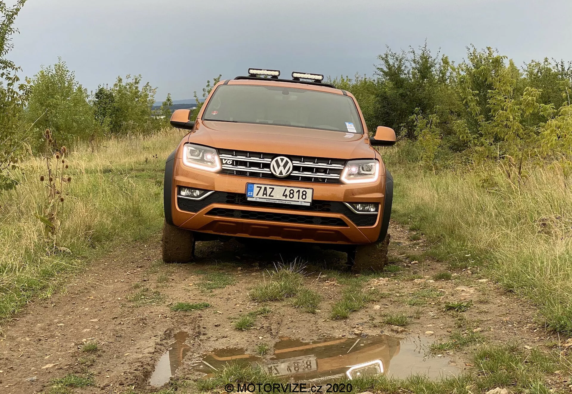 Widok z przodu na metaliczny pomarańczowy Volkswagen Amarok 2016 SUV na nieutwardzonym terenie z trawą i krzewami w tle. Pojazd jest skierowany w stronę kamery pod niewielkim kątem w dół, z płytką kałużą wody na pierwszym planie odbijającą obraz samochodu. Przednia krata ma w centrum logo Volkswagena, a maska pokazuje swoje kontury i linie wzornicze. Wyposażony w eleganckie reflektory LED i światła przeciwmgielne poniżej, srebrna osłona podwozia na dole sugeruje możliwości terenowe. Światła pomocnicze zamontowane są na bagażniku dachowym, co podkreśla jego solidny wygląd. Tablica rejestracyjna znajduje się centralnie na przednim zderzaku. Zdjęcie uchwyca pełną szerokość pojazdu, ukazując szczegóły związane z technologią, wzornictwem i adaptacjami terenowymi.