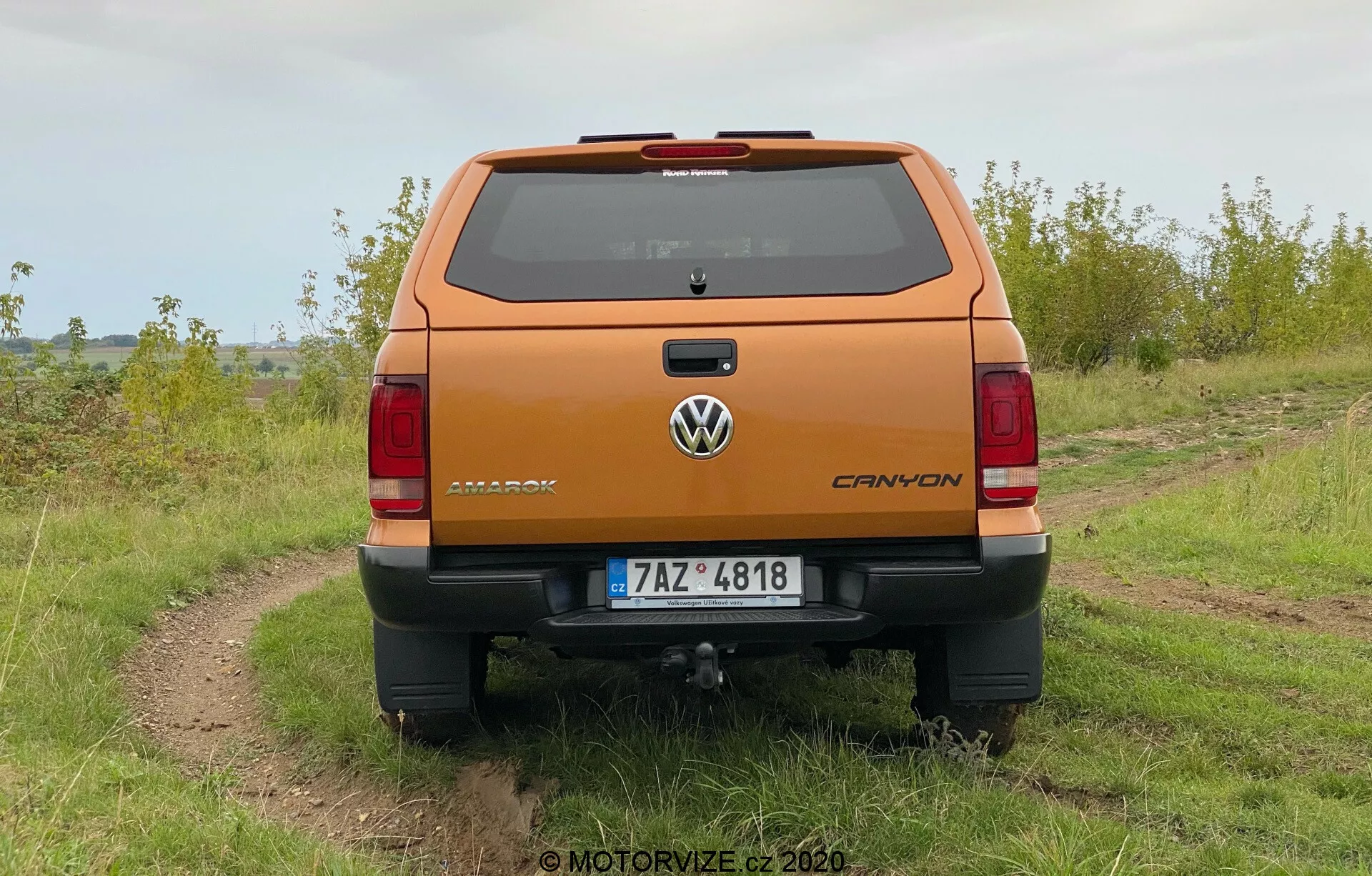 Tylny widok na metaliczno-pomarańczową Volkswagen Amarok 2016 pick-up zaparkowaną na zewnątrz na nierównej drodze szutrowej z trawą po obu stronach, pod pochmurnym niebem. Zdjęcie ukazuje wyraźną perspektywę tylnej części pojazdu, w tym logo Volkswagen, tabliczkę z nazwą Amarok i emblemat V6. Pojazd posiada tylni zderzak z wbudowanym stopniem, centralnie umieszczoną tablicę rejestracyjną i widoczny hak holowniczy. Widoczne są także pionowo zorientowane światła tylnie oraz trzecie światło stopu na linii dachu nad tylną szybą, jak również linie rozmrażacza.