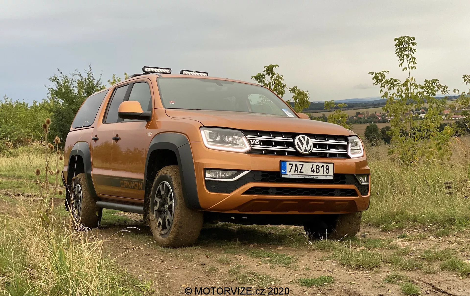Obraz przedstawia nowoczesne SUV, model Volkswagen Amarok z 2016 roku, zaparkowane na zewnątrz na nierównym, ziemistym terenie, demonstrując swoje zdolności terenowe. Widok z niskiego przedniego kąta ukazuje przednią ćwiartkę pojazdu pokrytą błotnistymi resztkami po niedawnym użyciu w terenie. Szeroka przednia kratka z charakterystycznym emblematem VW jest otoczona prostokątnymi reflektorami z diodami LED do jazdy dziennej. Poniżej kratki znajduje się solidny przedni zderzak z srebrną płytą ochronną zaprojektowaną do ochrony podwozia. Konturowana maska dodaje SUV-owi muskularnego wyglądu. Widoczne są relingi dachowe, wskazując na dodatkową zdolność do przewozu ładunku. Koła, częściowo zasłonięte błotem, mają głęboki bieżnik do lepszej trakcji na trudnym terenie. Widoczny jest wyraźny łuk nad przednim kołem i stopień boczny ułatwiający wejście i wyjście z pojazdu. Widoczne są także oświetlenie dachowe i lusterka boczne. Samochód jest umieszczony na tle roślinności i pagórkowatego terenu pod pochmurnym niebem, podkreślając swoje zdolności terenowe i atrakcyjność na świeżym powietrzu. Lewa kierownica i widoczny numer rejestracyjny sugerują kraj, w którym jeździ się po prawej stronie drogi. Ten kadr podkreśla design pojazdu, jego terenowe cechy i zaawansowane technologie oświetleniowe.