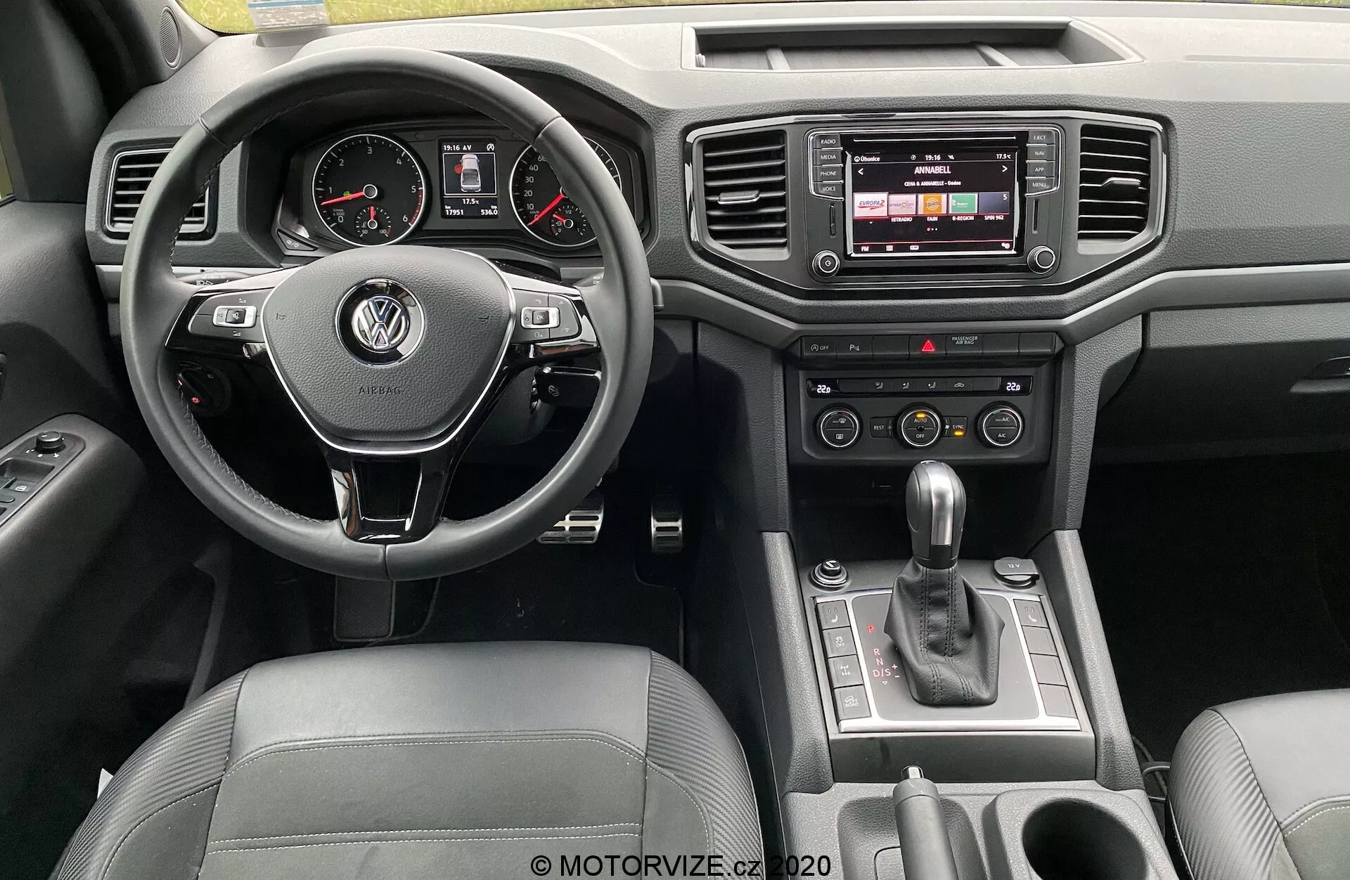 Widok deski rozdzielczej Volkswagena Amarok 2016 z perspektywy kierowcy. Centralnie umieszczona wielofunkcyjna kierownica obszyta skórą z logo VW, analogowymi miernikami i cyfrowym wyświetlaczem na desce rozdzielczej. Konsola centralna wyposażona jest w system infotainment z ekranem dotykowym, przyciskami do regulacji klimatyzacji i dźwignią zmiany biegów automatycznej. Luksusowe detale obejmują wykończenie ze skóry i chromu, widoczne są również przyciski na drzwiach.