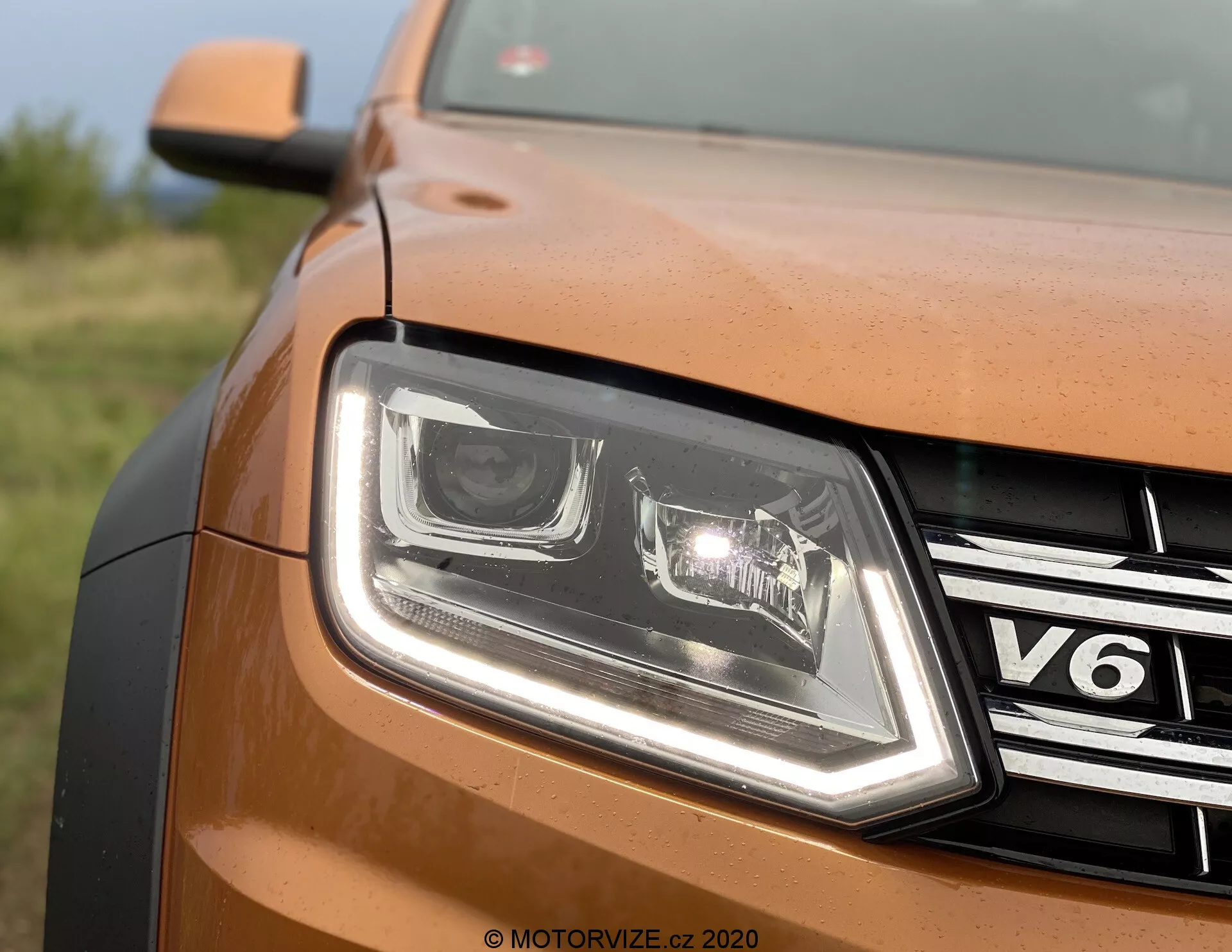 Zbliżenie częściowego przedniego widoku z lewej strony brązowego lub miedzianego VW Nutzfahrzeuge Amarok 2016. Obraz pokazuje projekty reflektorów LED z wyraźnymi światłami do jazdy dziennej, chromowaną kratkę z odznaką 'V6', metaliczny lakier panelu nadwozia, obrysowaną krawędź maski oraz znak wodny 'MOTORVIZE.CZ 2020', co sugeruje pochodzenie z czeskiego serwisu recenzji samochodowych.