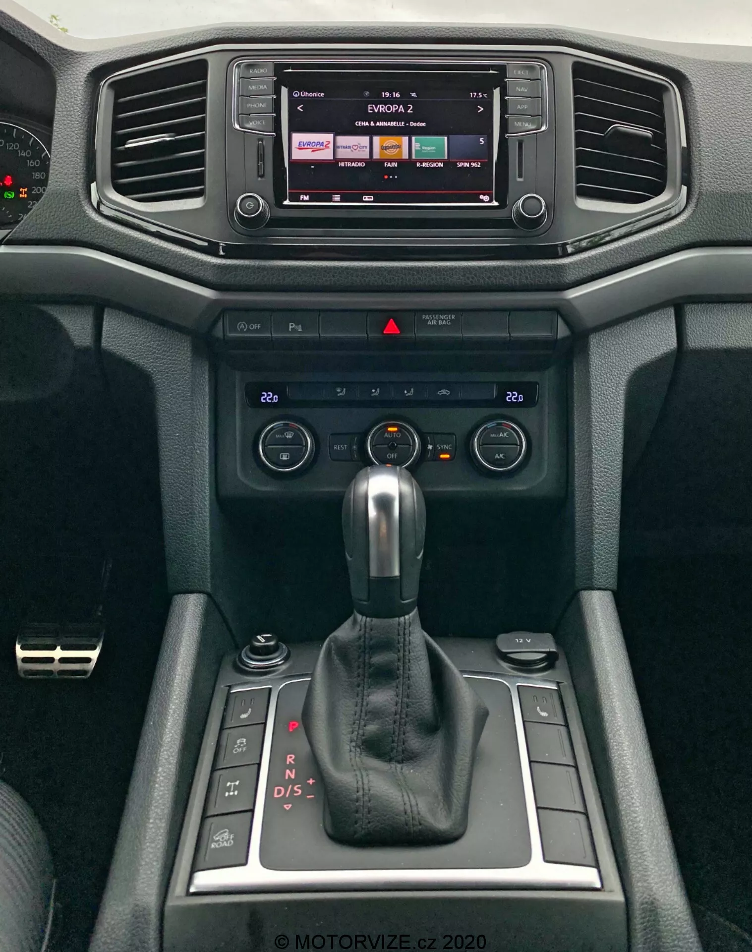 Zbliżenie na nowoczesną konsolę centralną VW Amarok 2016, z wyeksponowanym systemem infotainment i dźwignią zmiany biegów. Wyświetlacz infotainment sugeruje możliwości dotykowe, prawdopodobnie pokazując wyjście radiowe lub medialne. Poniżej znajdują się poziome nawiewy otoczone miękkimi materiałami o teksturowanym wzornictwie. Poniżej nawiewów umieszczono pokrętła i przyciski do sterowania klimatyzacją, umożliwiające regulację temperatury i przepływu powietrza. Na środku dominuje automatyczna dźwignia zmiany biegów w trybie 'P', wskazując na automatyczną skrzynię biegów z możliwością ręcznego zmieniania biegów. Po bokach dźwigni zmiany biegów znajdują się małe schowki oraz obrotowe pokrętło, prawdopodobnie do sterowania infotainment lub systemami trybów jazdy. Wnętrze samochodu podkreśla funkcjonalność i komfort.
