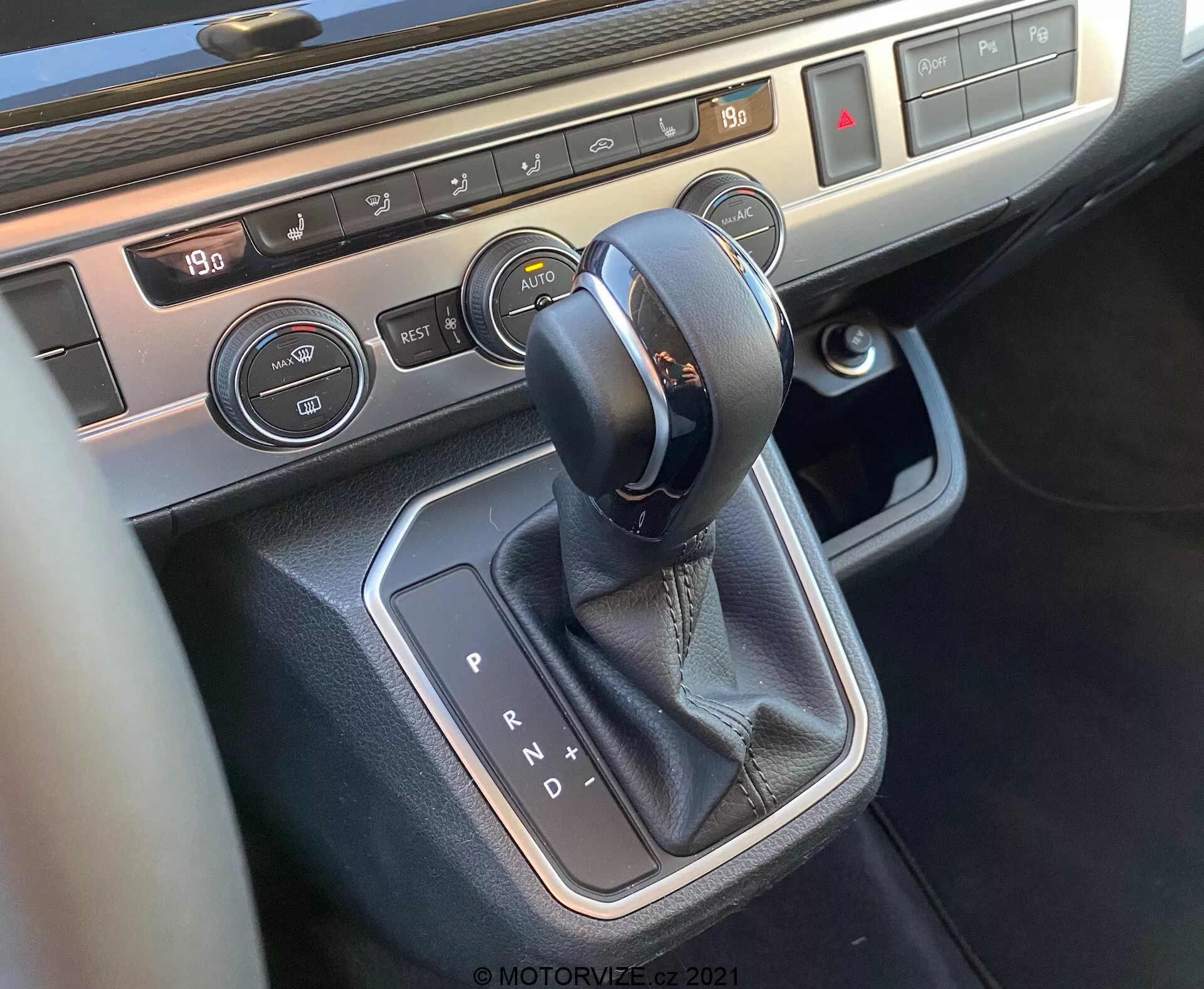 Zbliżenie konsoli środkowej VW Nutzfahrzeuge T6-1 Multivan 2019, skupiające się na obszarze dźwigni zmiany biegów. Obraz przedstawia automatyczny selektor skrzyni biegów z przeszyciem skórzanym, otoczony metalową obudową, przyciski sterowania klimatyzacją z wyświetlaczem temperatury oraz elementy sterowania ogrzewaniem siedzeń, podkreślając nowoczesną technologię wnętrza i materiały premium pojazdu.