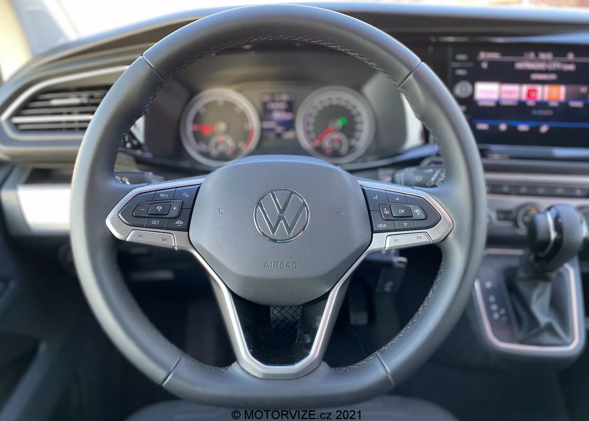 Widok wnętrza samochodu VW Nutzfahrzeuge Transporter 2019 (T6-1 Multivan) z perspektywy kierowcy. Szczegółowy widok kierownicy z logo Volkswagena i przyciskami sterującymi dla różnych funkcji, takich jak tempomat i nawigacja systemu multimedialnego. Za kierownicą znajduje się zespół wskaźników z analogowym prędkościomierzem i obrotomierzem oraz cyfrowym wyświetlaczem między nimi. Częściowo widoczna konsola środkowa z dużym ekranem dotykowym infotainment, wraz z eleganckim wzornictwem deski rozdzielczej z poziomym otworem wentylacyjnym. Dźwignia zmiany biegów po prawej stronie wskazuje na automatyczną skrzynię biegów. Zdjęcie podkreśla funkcje użytkowe z małą głębią ostrości, koncentrując się głównie na kierownicy.