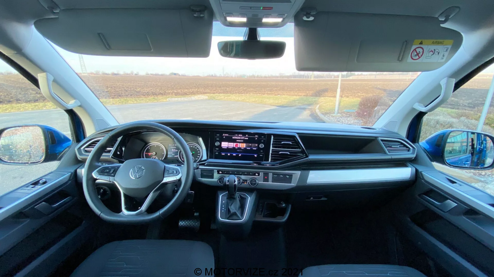 Wnętrze VW Nutzfahrzeuge Transporter 2019 (T6.1 Multivan), uchwycone z tylnego siedzenia w kierunku przedniej deski rozdzielczej. Zdjęcie zrobiono w ciągu dnia, kiedy naturalne światło wpada przez przednią szybę, zapewniając szczegółowy widok przestrzeni kabiny, przednich siedzeń, kierownicy z wbudowanymi przyciskami, centralnego ekranu dotykowego systemu informacyjno-rozrywkowego, przycisków i pokręteł klimatyzacji, automatycznego lewarka zmiany biegów, schowków, portów łączności, przycisków drzwi i lusterek, funkcji bezpieczeństwa oraz wygodnego projektu siedzeń.