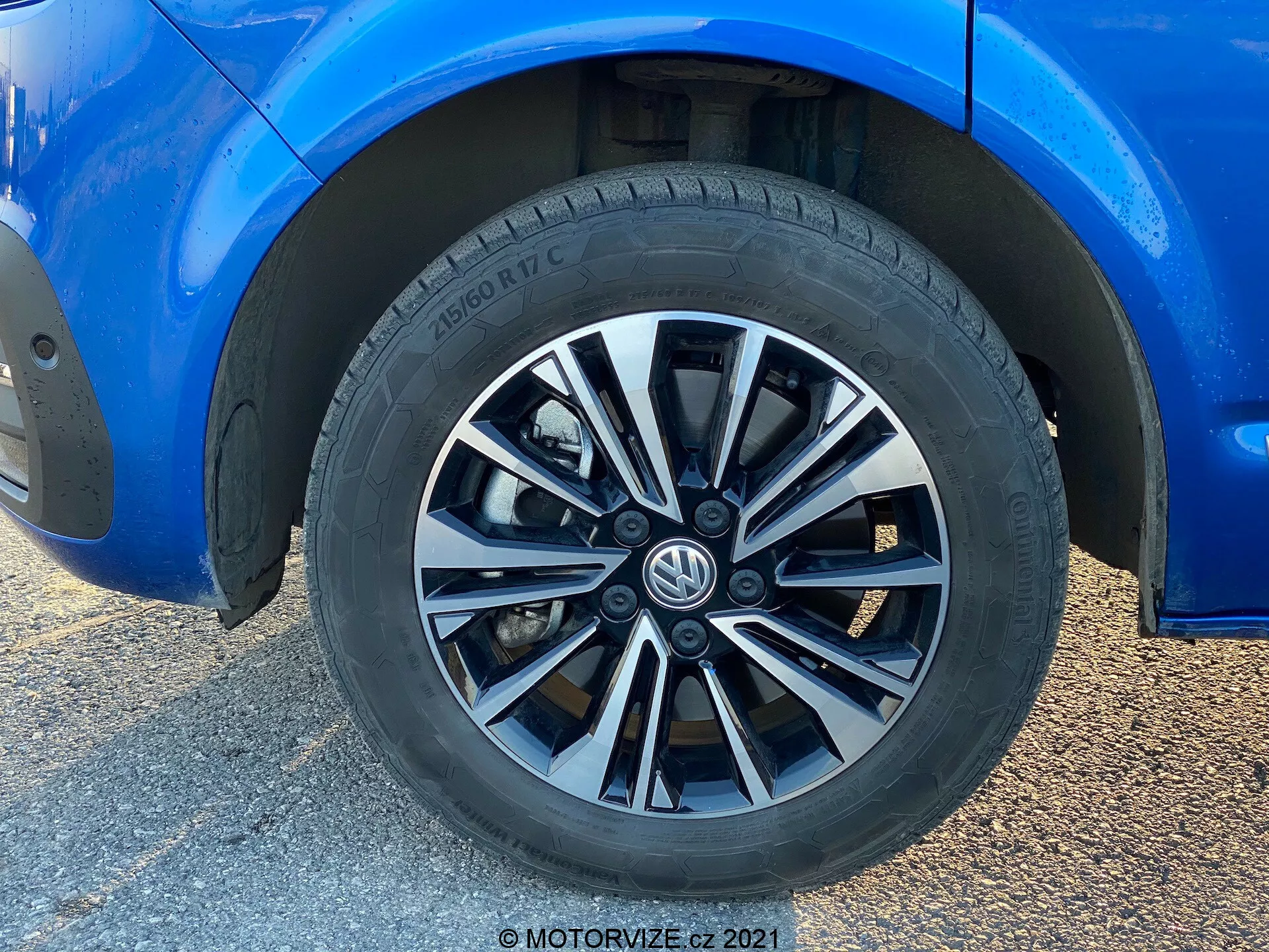 Zbliżenie na koło Volkswagen T6-1 Multivan 2019. Ten pełnokadrowy strzał przedstawia felgę wieloramienną z naprzemiennymi srebrnymi i ciemnymi wykończeniami. Opona marki Hankook pokazuje wzór bieżnika przeznaczony do wydajności drogowej i częściowe specyfikacje opony. Otaczająca niebieska karoseria, w doskonałym stanie, pokazuje refleksyjny połysk. Wskazuje na umiarkowany prześwit i częściowo widoczne standardowe zaciski hamulcowe i tarcze. Naturalne światło podkreśla kompozycję skoncentrowaną na kole, z rozmytym tłem w wyniku niskokątowego zbliżenia.