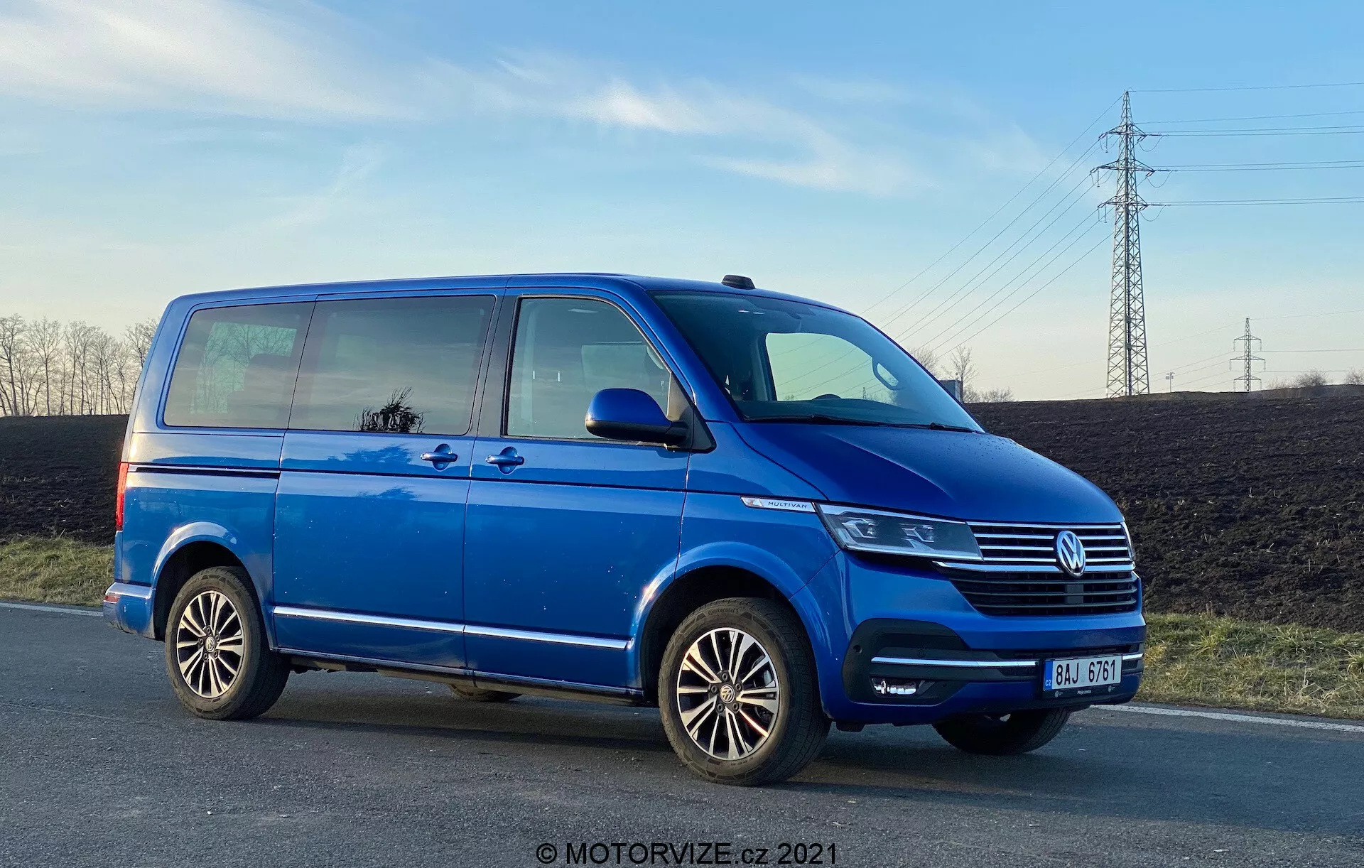 Niebieski Volkswagen Transporter T6, pokazany z profilu bocznego z niskiego kąta, podkreślający jego długość i wysokość. Obraz pokazuje szczegóły takie jak przesuwane drzwi pasażera, przednie drzwi pasażera, tylne koło po stronie pasażera, wieloramienne felgi aluminiowe oraz częściowe widoki reflektora, kraty Volkswagena z emblematem i zderzaka przedniego. Zdjęcie, zrobione późnym popołudniem, pokazuje prześwit od ziemi i odbija niebo oraz otoczenie w oknach, z czystym niebem i liniami wysokiego napięcia w tle.