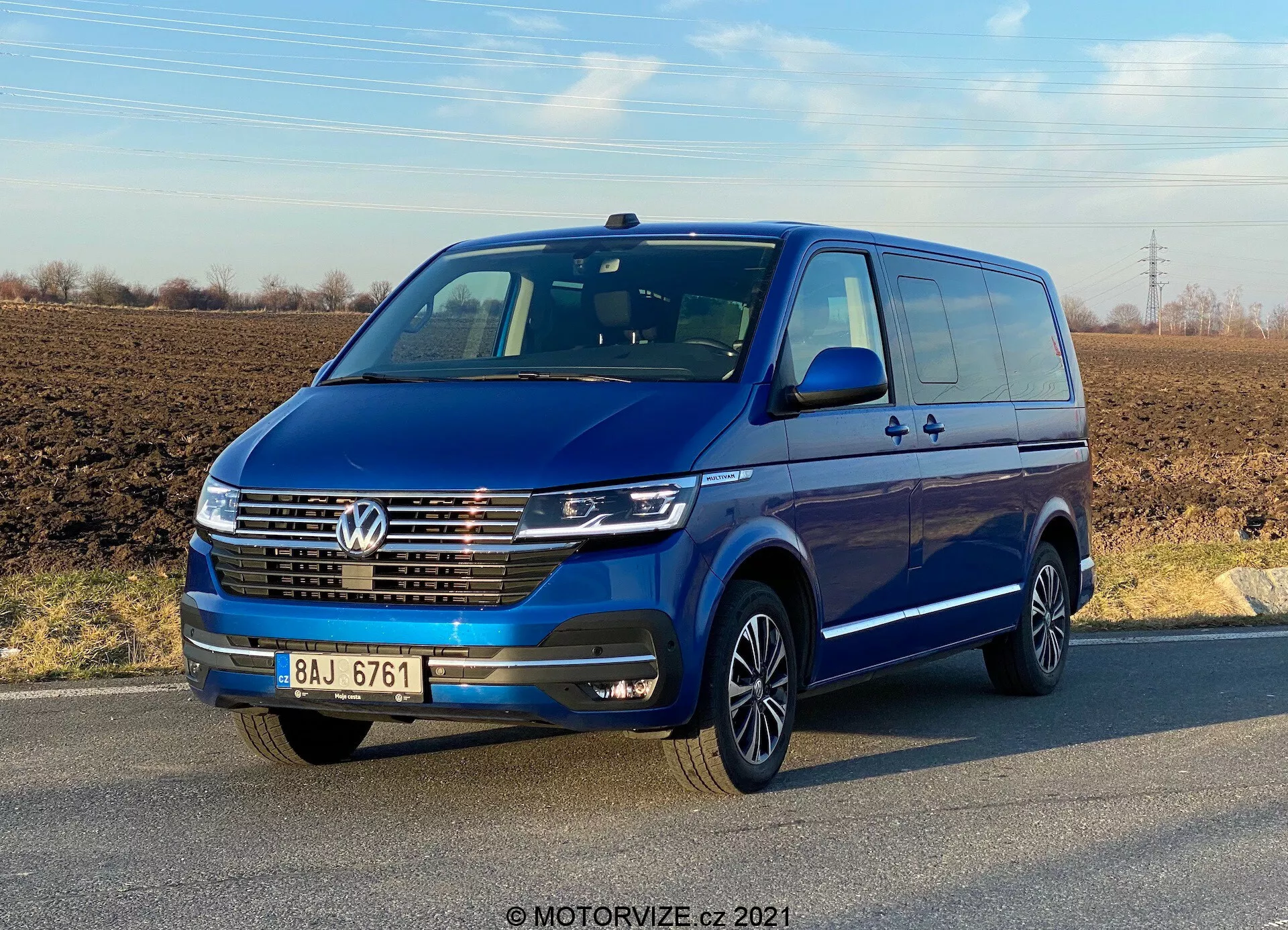 Widok trzy czwarte z przodu na niebieskiego Volkswagena Transportera T6.1 Multivana 2019 pod czystym niebem. Pojazd posiada znaczek Volkswagena na grillu, światła do jazdy dziennej LED, chromowane listwy na przednim grillu oraz wieloramienne felgi aluminiowe. Lusterka i zderzaki w kolorze nadwozia sugerują model o wysokiej specyfikacji. Widać drzwi i klamki, w tym tylne drzwi przesuwne, co wskazuje na możliwość przewozu wielu pasażerów lub ładunku.
