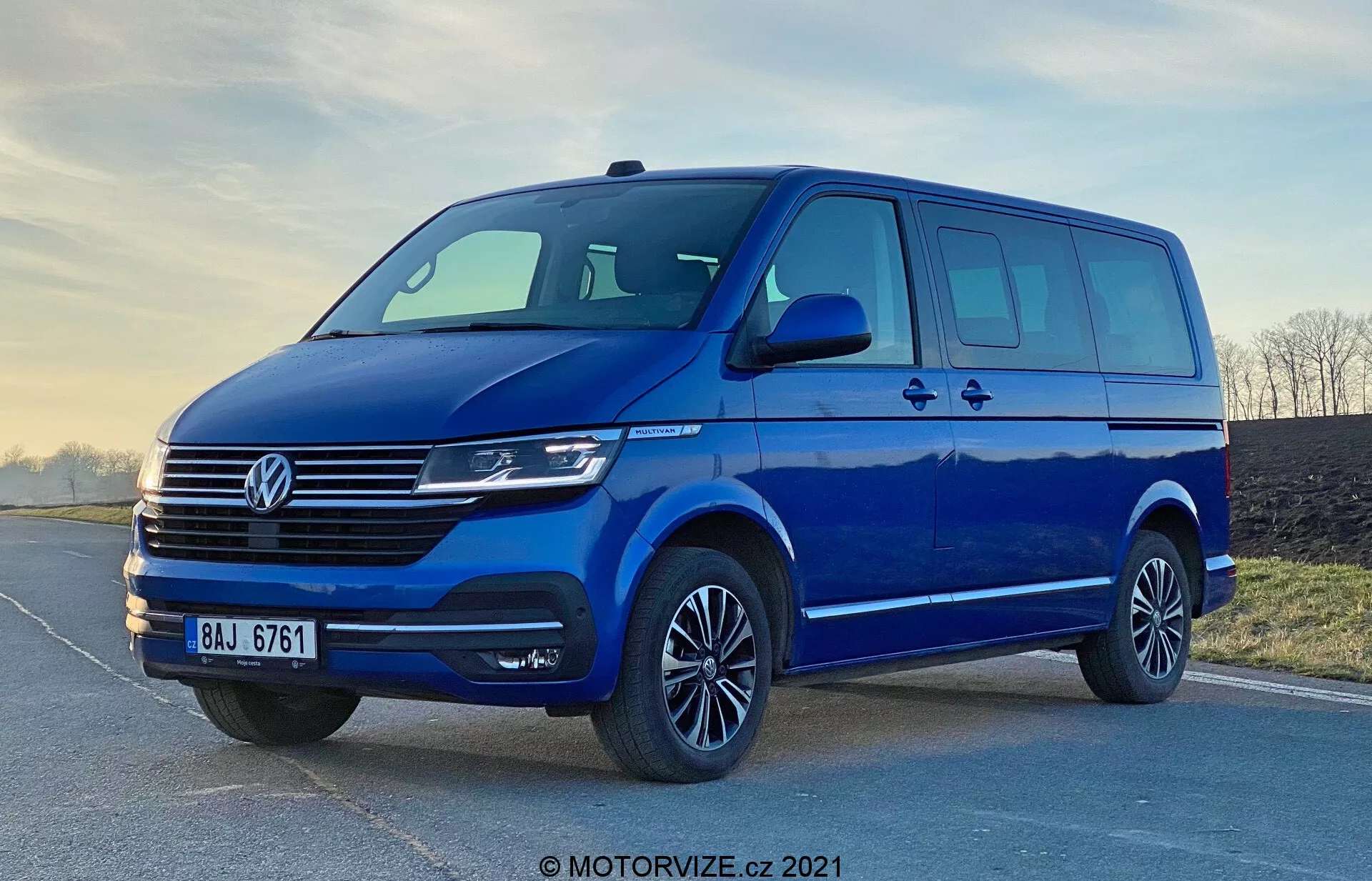 Obraz niebieskiego Volkswagena Transportera T6.1 Multivan z 2019 roku, uchwycony z niskiego kąta, aby podkreślić jego posturę i wielkość. Widok z przodu z trzech czwartych ukazuje front i bok pojazdu, w tym charakterystyczny dla Volkswagena grill z poziomymi liniami, centralny emblemat, nowoczesne reflektory prawdopodobnie z technologią LED lub Xenon oraz elegancki przedni zderzak z szerokim wlotem powietrza. Aerodynamiczne lusterka boczne z zintegrowanymi kierunkowskazami, duża powierzchnia okien z bocznymi drzwiami przesuwnymi, felgi aluminiowe z oponami o niskim profilu oraz miękkie oświetlenie podkreślają praktyczność i estetykę furgonu. Znak wodny 'MOTORVIZE.cz 2021' sugeruje, że obraz pochodzi z 2021 roku.