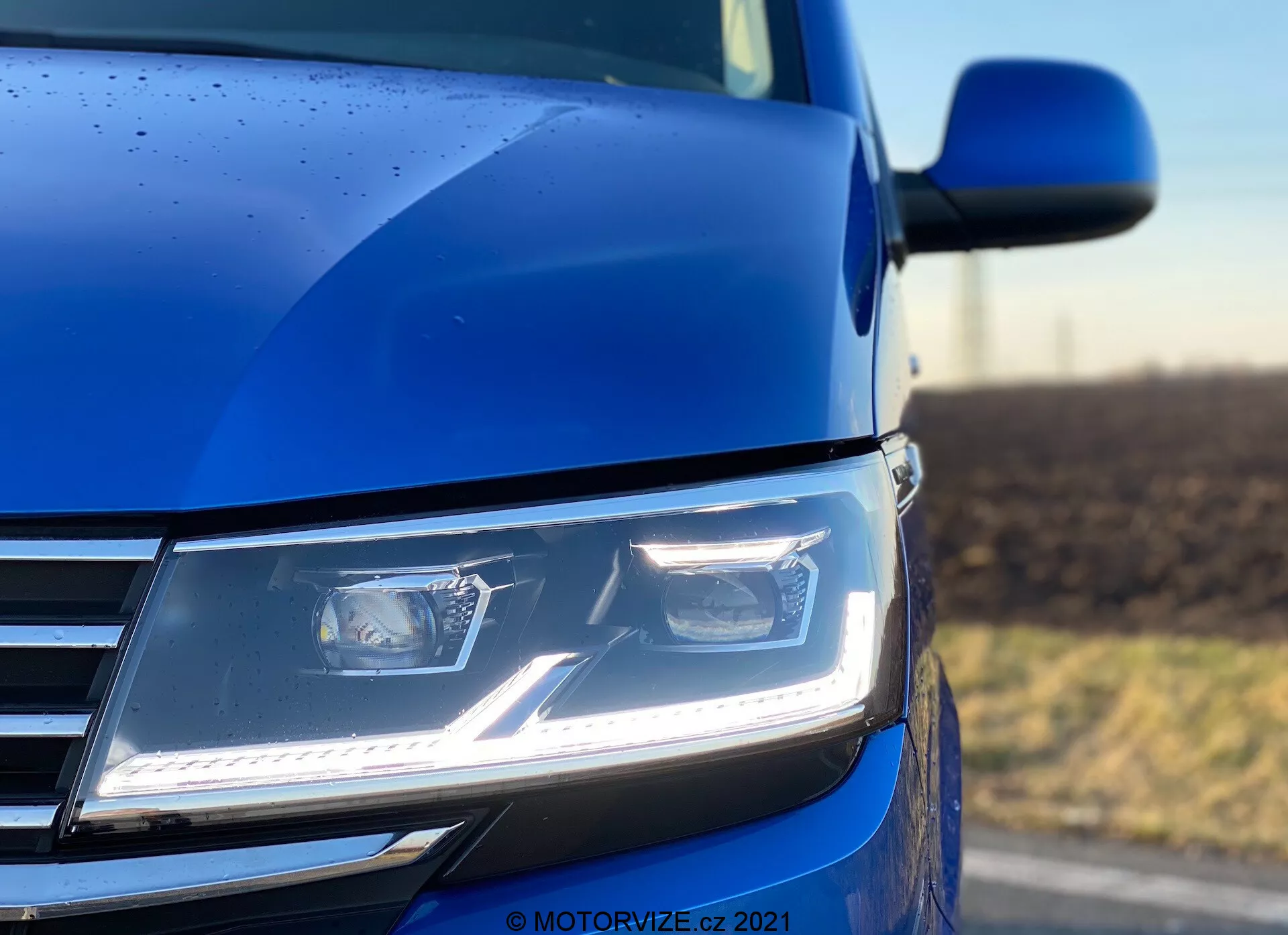 Zbliżenie przedniej lewej sekcji niebieskiego VW Nutzfahrzeuge Transporter 2019 (T6.1 Multivan) z niskiego kąta, z nowoczesnymi, kątowymi reflektorami LED z światłami dziennymi, częścią chromowanej atrapy chłodnicy oraz fragmentem konturu maski i lusterkiem bocznym. Zdjęcie wykonane przy małej głębi ostrości, podkreślające reflektor i otaczającą karoserię, uwydatniając zaawansowaną technologię oświetlenia, nowoczesny design i jakość lakieru, uzupełnione o krople deszczu wskazujące na szczegółowe pokrycie zewnętrzne.